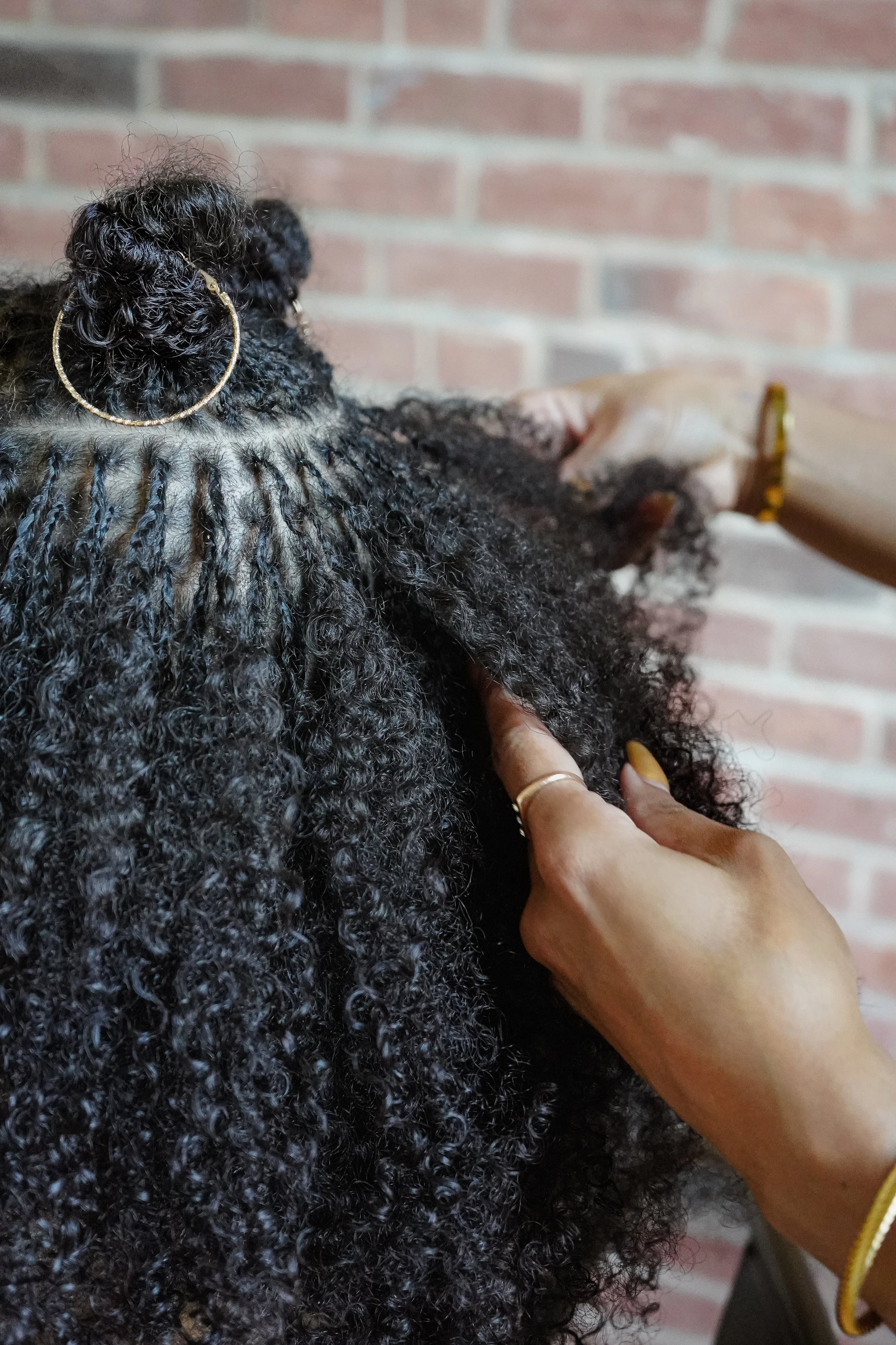 A person with dark, curly hair styling or braiding another person's hair against a brick wall background.