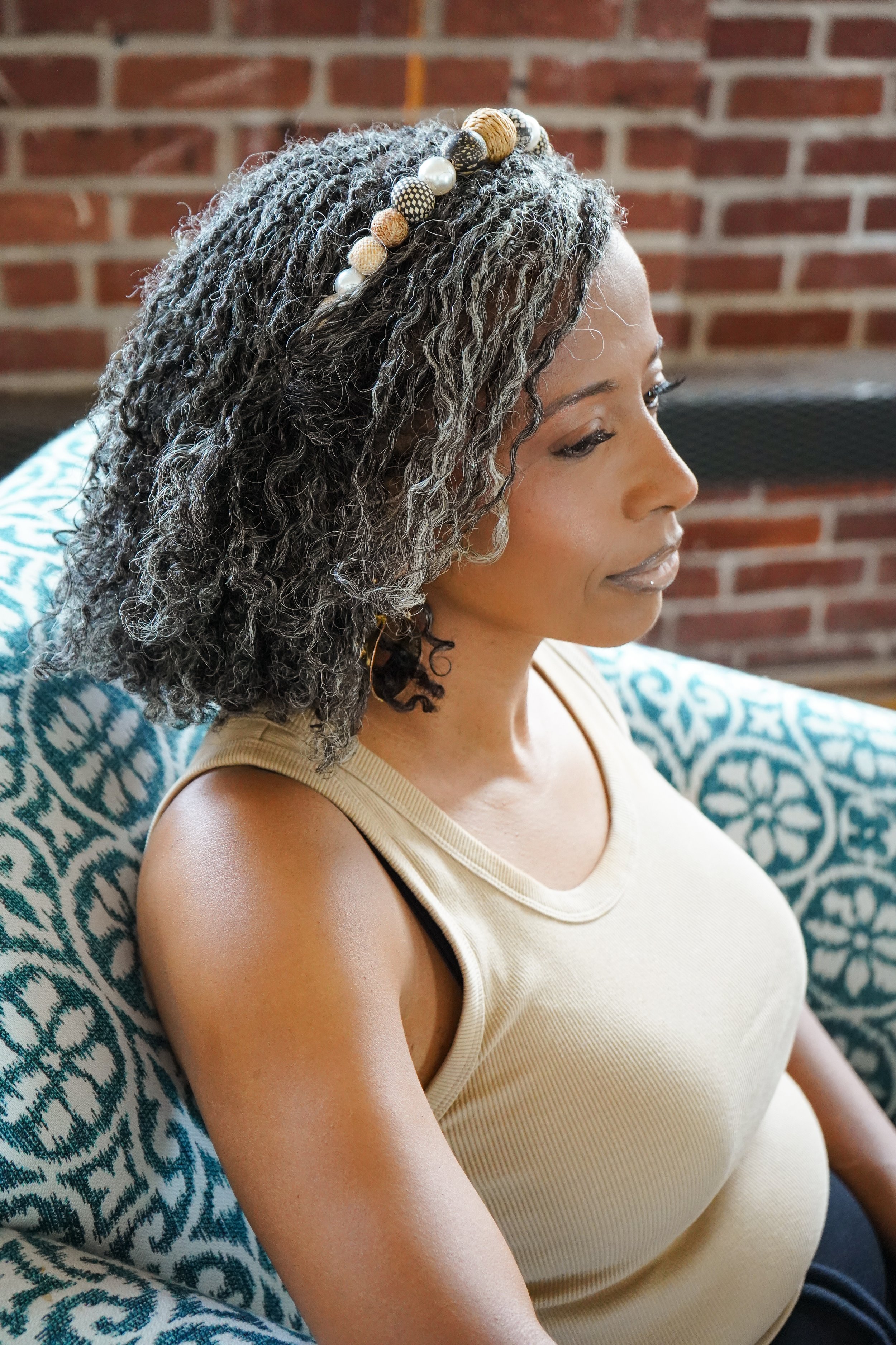Side view of a woman with gray, newly installed microlocs wearing a beaded headband, beige tank top, sitting on a patterned blue and white couch against a brick wall background.