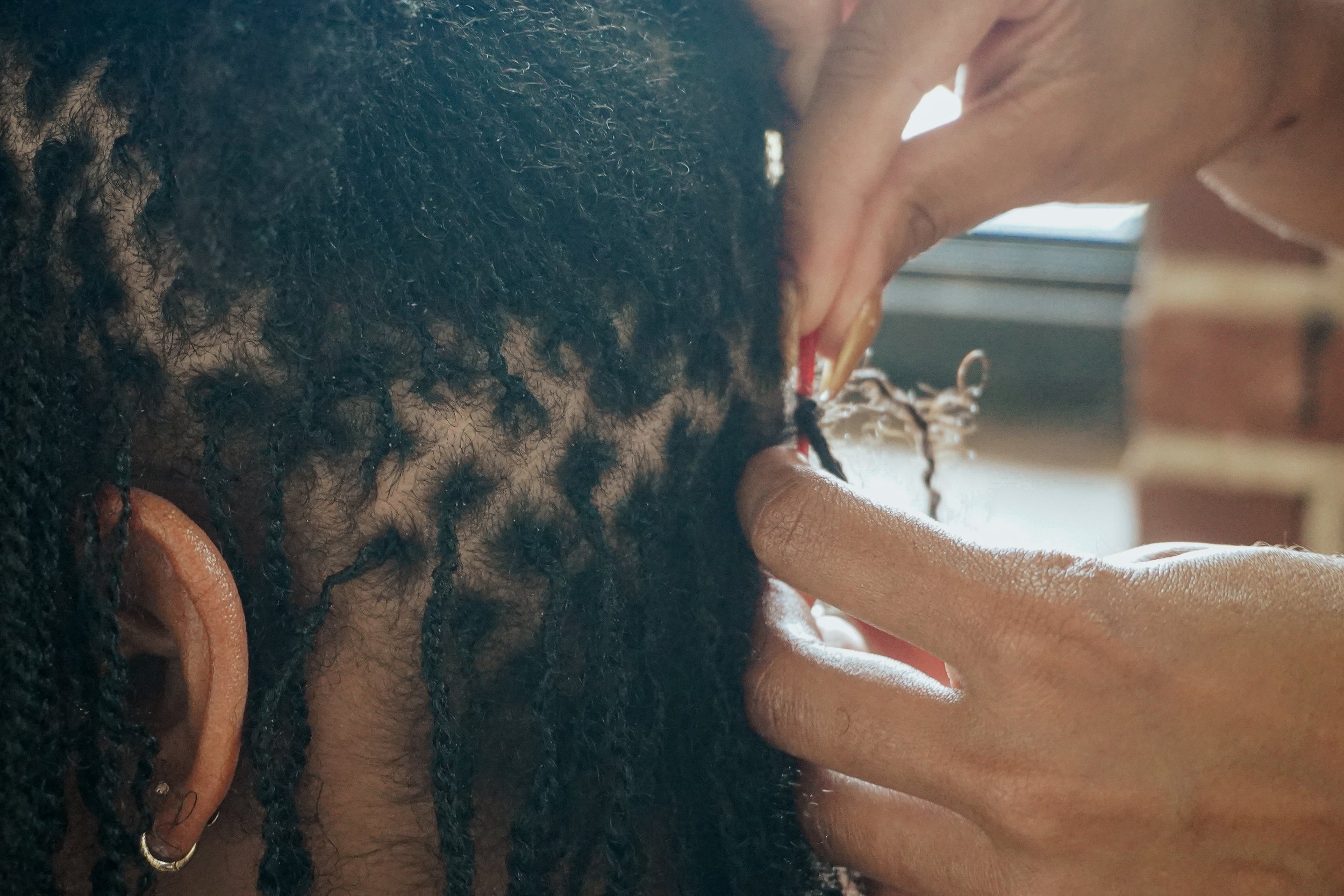 A person is holding a needle to retwist small, natural black microlocs with their right hand, retwisting.
