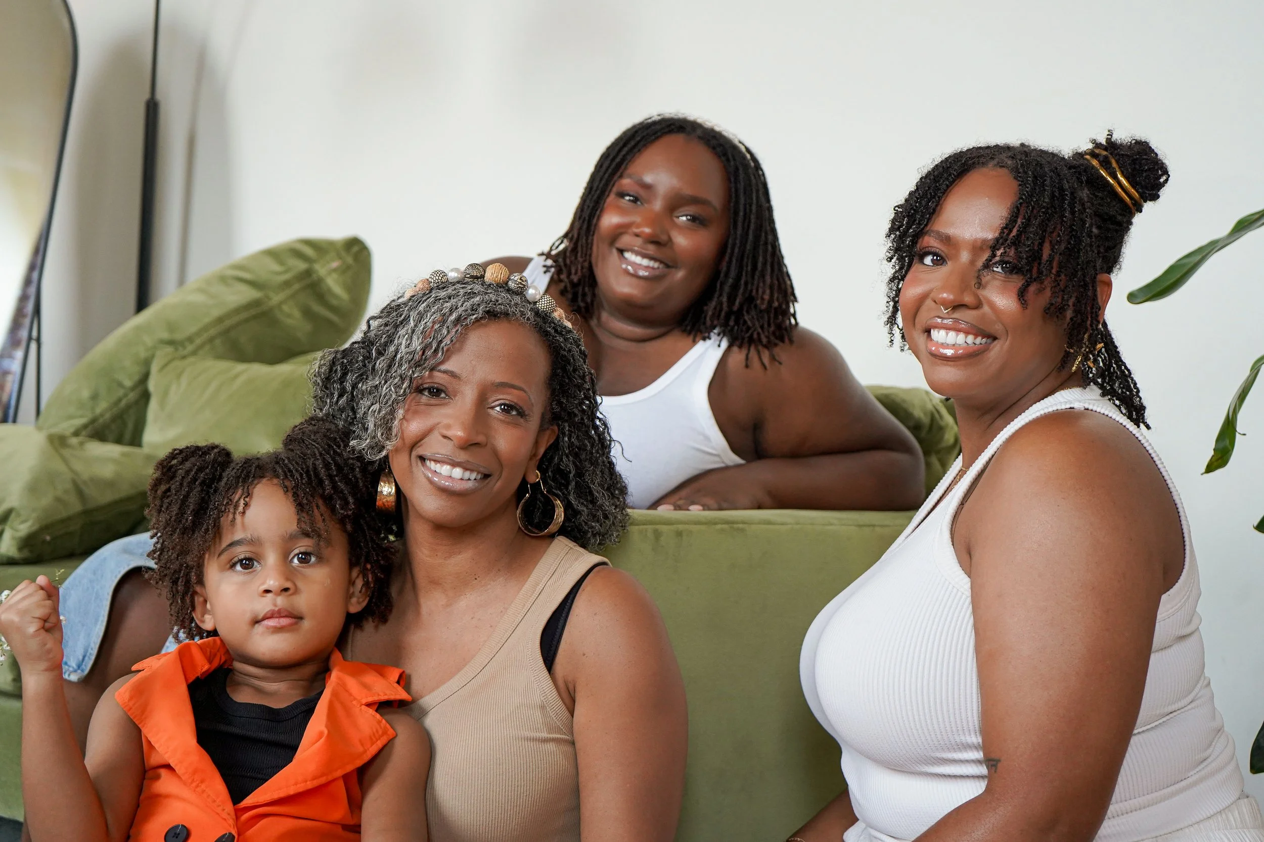Four women and one young girl sitting on a green couch, smiling in a living room.