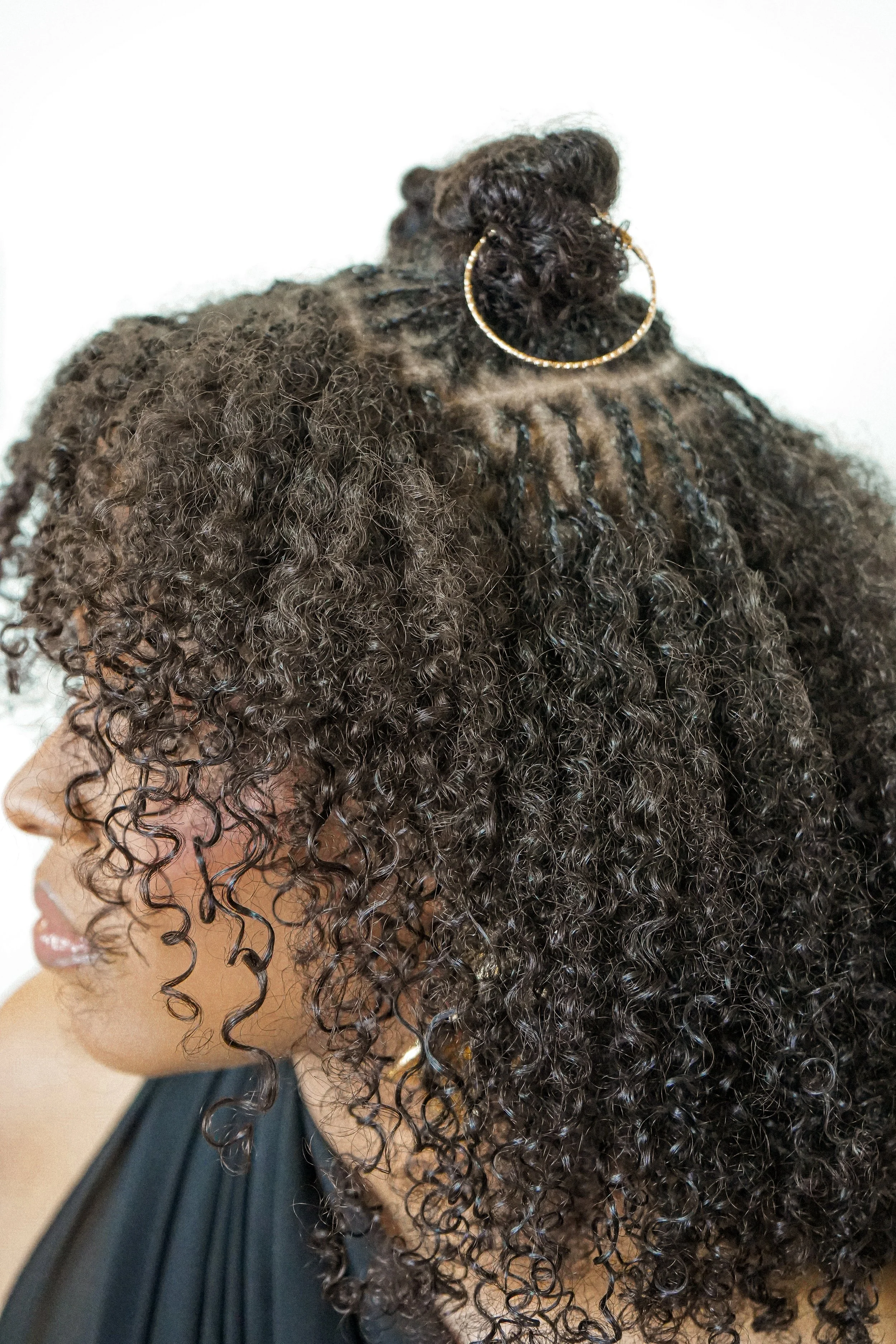 Close-up of a woman with curly hair and root braids styled in an updo, wearing large hoop earrings, and a black top with ruffled details on the shoulder, against a white background.