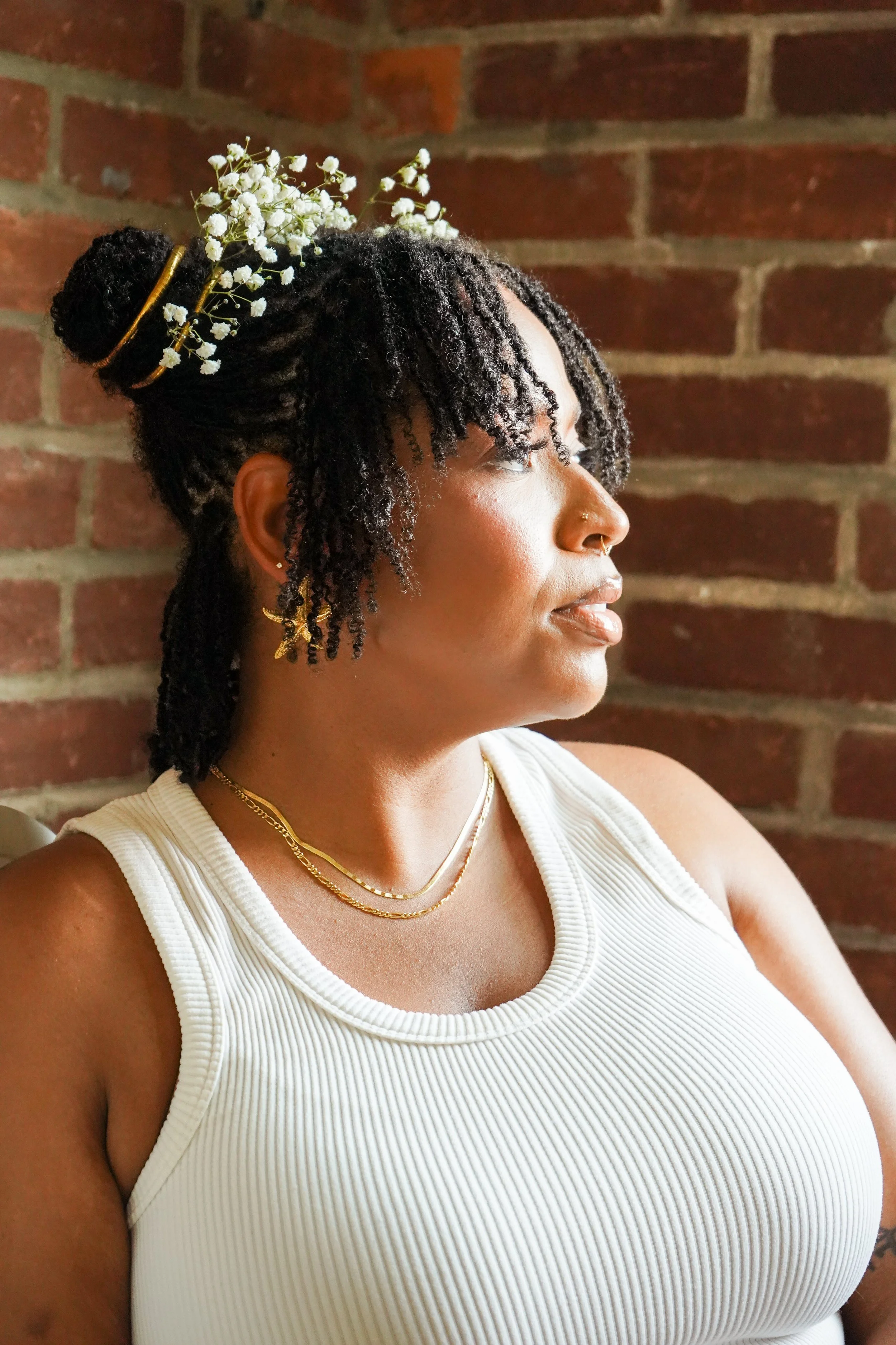 A woman with styled microlocs and a floral hair accessory looking to her right, standing in front of a brick wall.