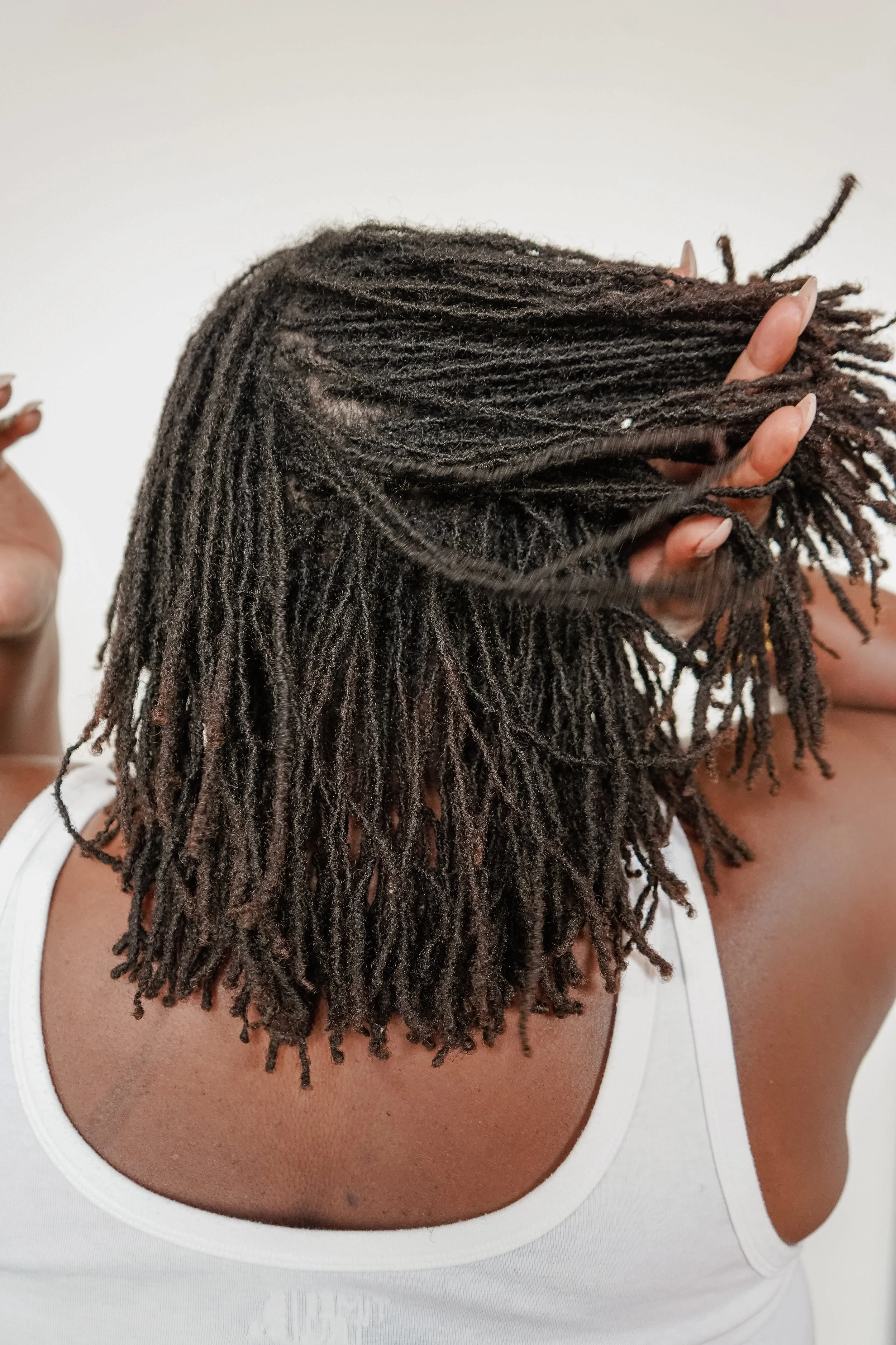 A person with long, thick dreadlocks wearing a white top, holding their hair with one hand, against a plain white background.