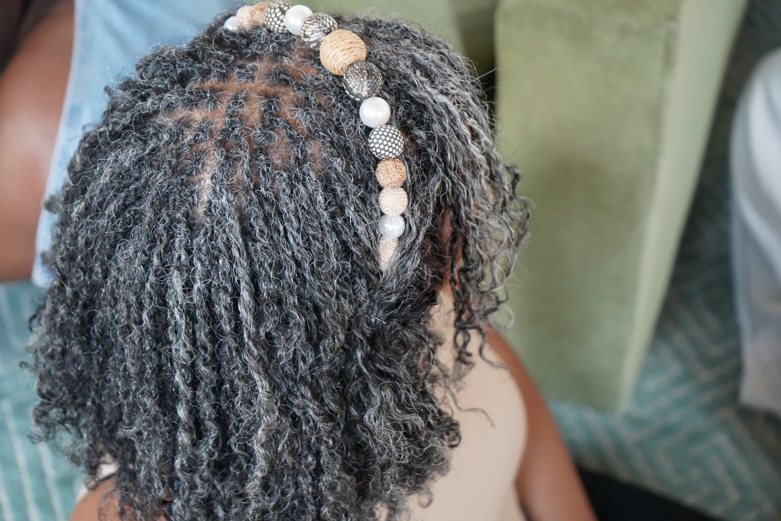 Close-up of a person's head with curly gray and black hair, adorned with a beaded hair accessory featuring various patterned beads.