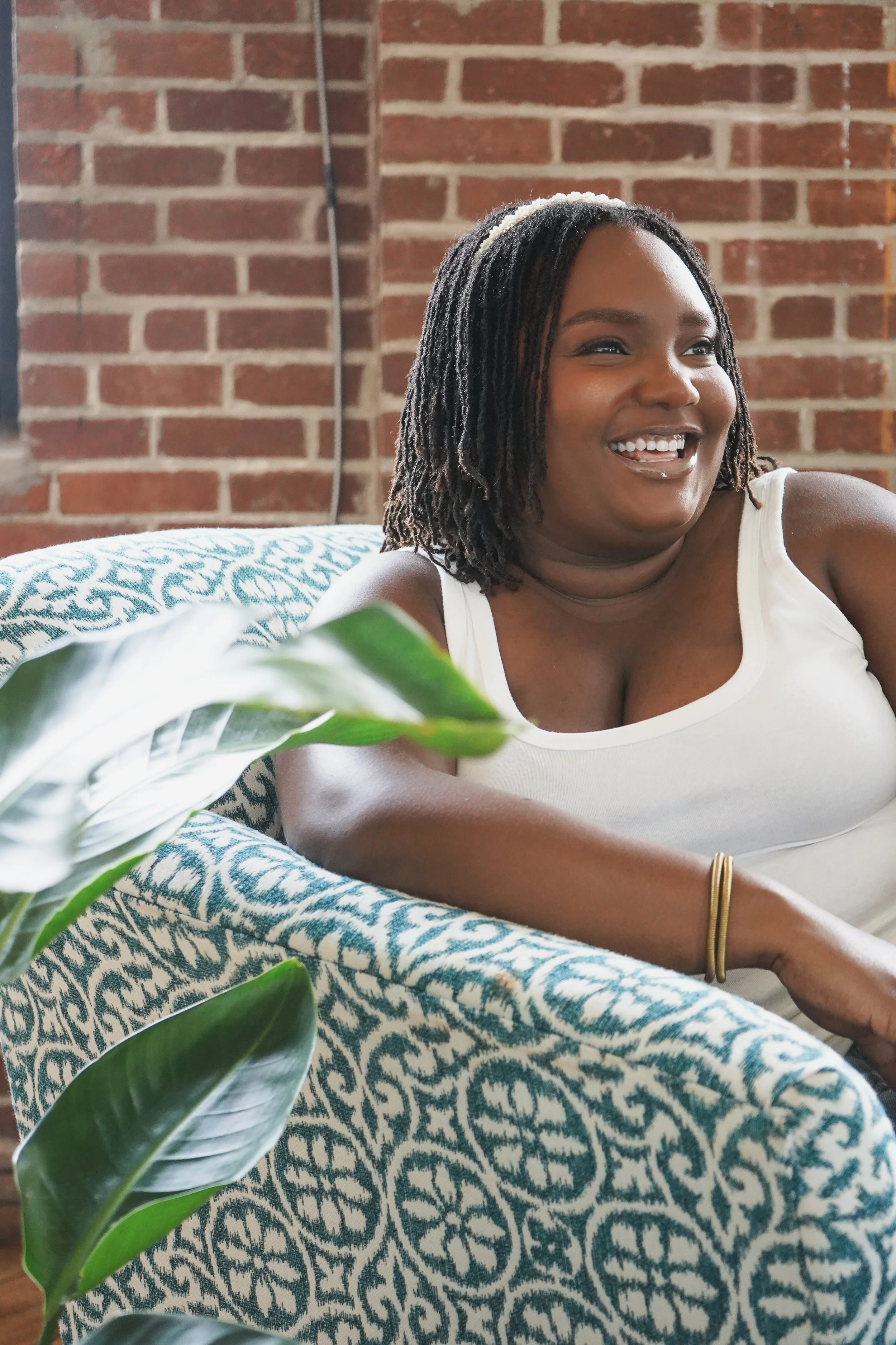 A woman with microlocs and a white tank top sitting on a patterned blue and white couch, smiling while looking to her right, against a brick wall background and a green plant in the foreground.