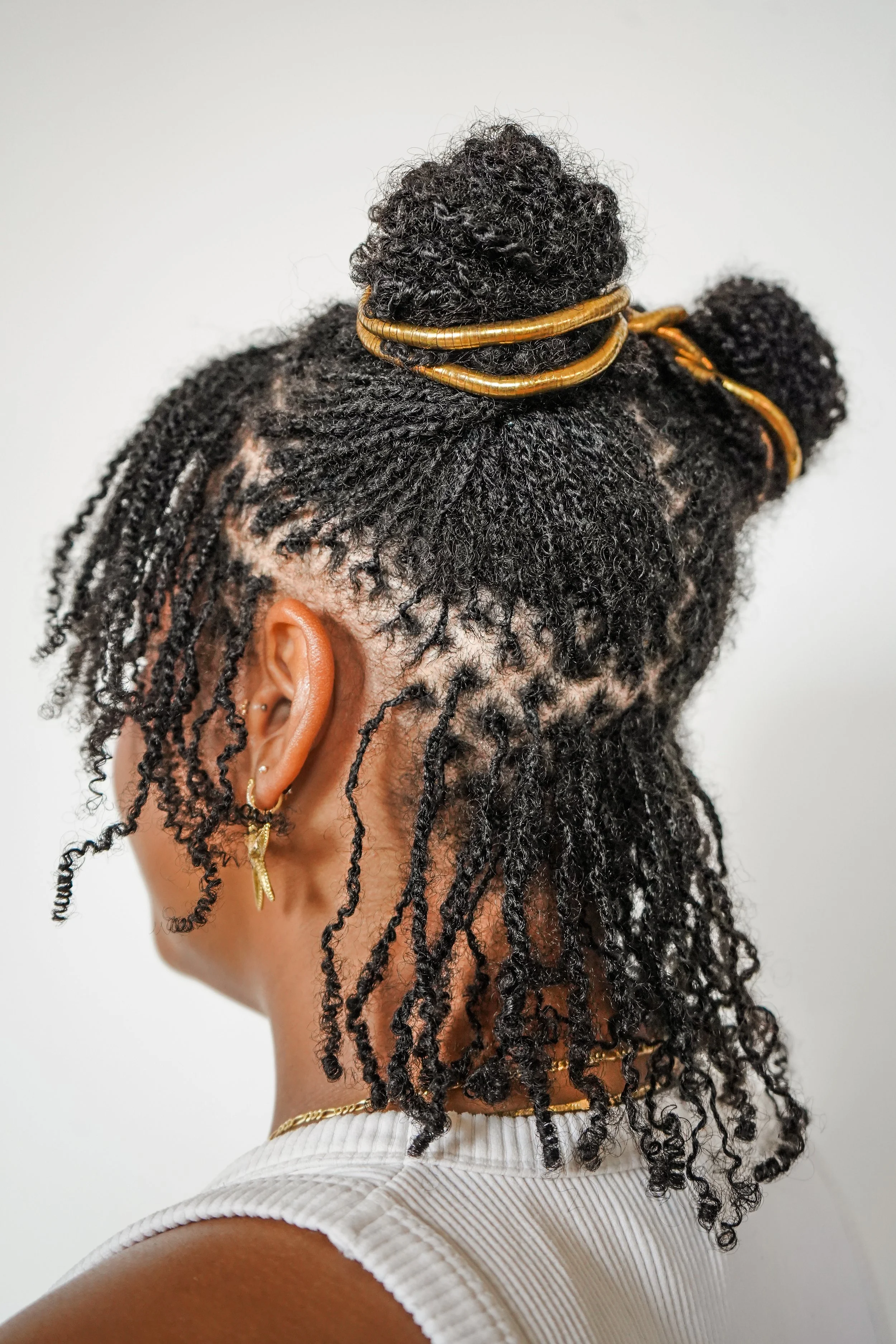 A woman with natural black twisted and coiled hair styled with gold accessories, wearing earrings, a necklace, and a white sleeveless top.