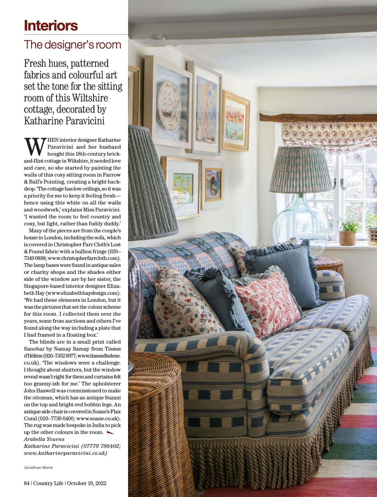 Cozy living room with a floral and striped sofa adorned with various patterned cushions. The room features a large glass lamp with a patterned lampshade, framed artwork on the wall, and a window with floral curtains. Potted plants and a small side ta