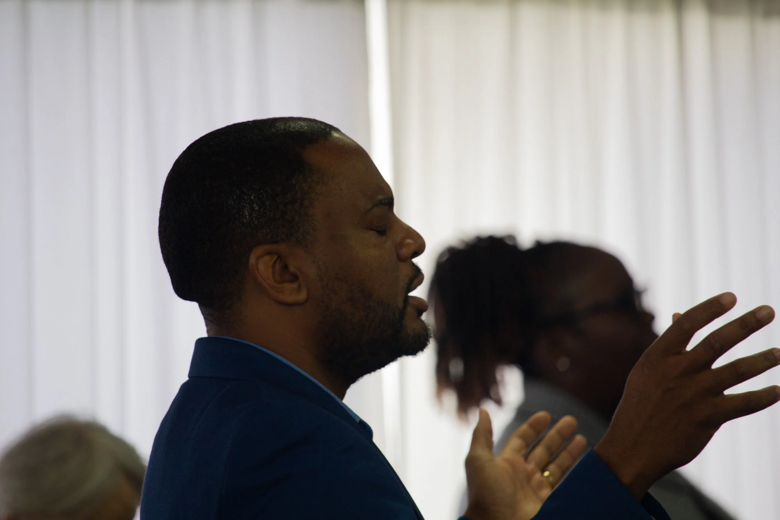 Man with short hair and beard wearing a blue suit, eyes closed, hands raised, participating in a prayer or meditation session in a room with white curtains. Pictured at a Greaterlife Church Sunday Service.