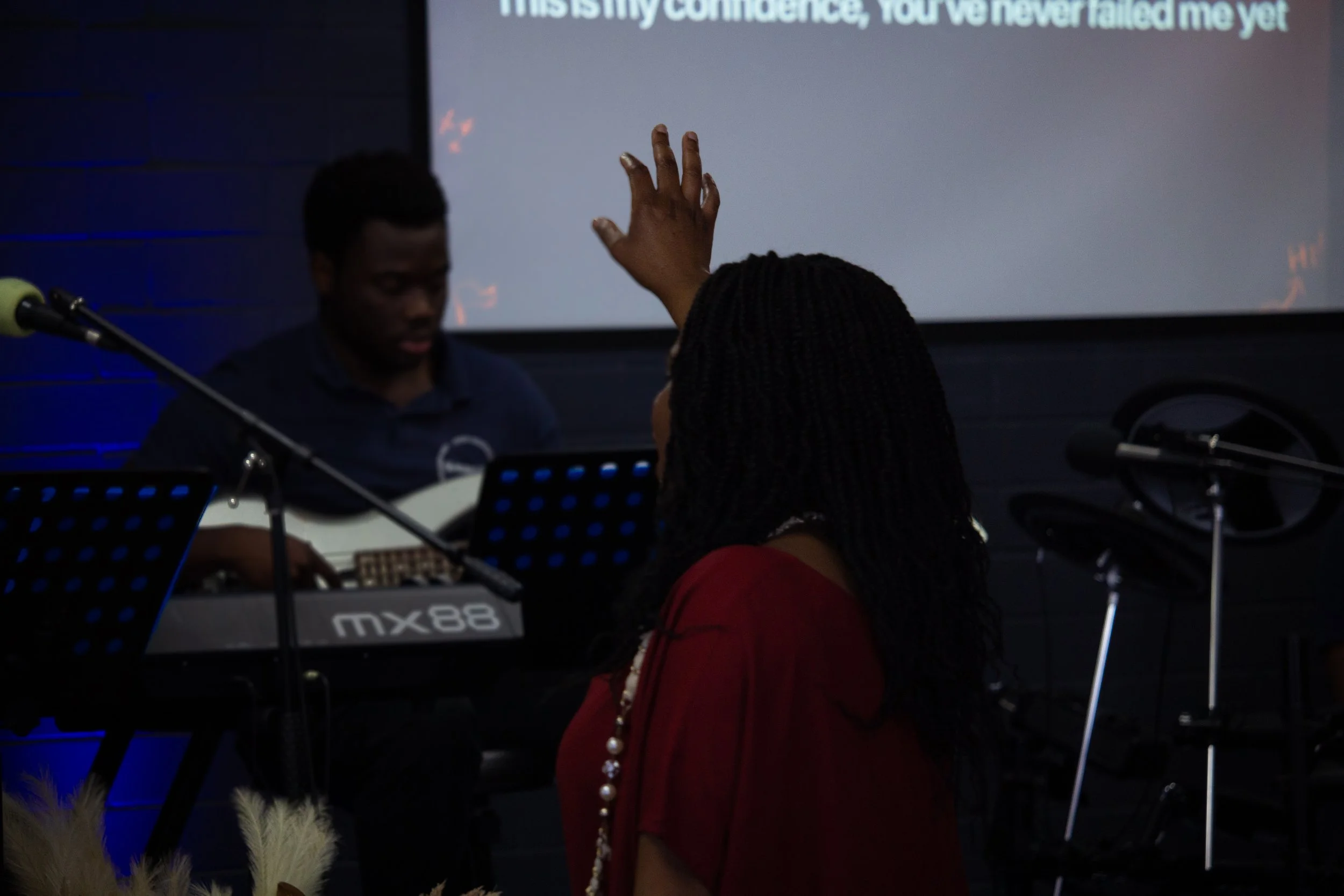 Pastor Yemu Phiri, raising her hand during a performance or event, with a man playing a keyboard and a guitarist in the background in a dark room with a large screen displaying text. Pictured at a Greaterlife Church Sunday Service.