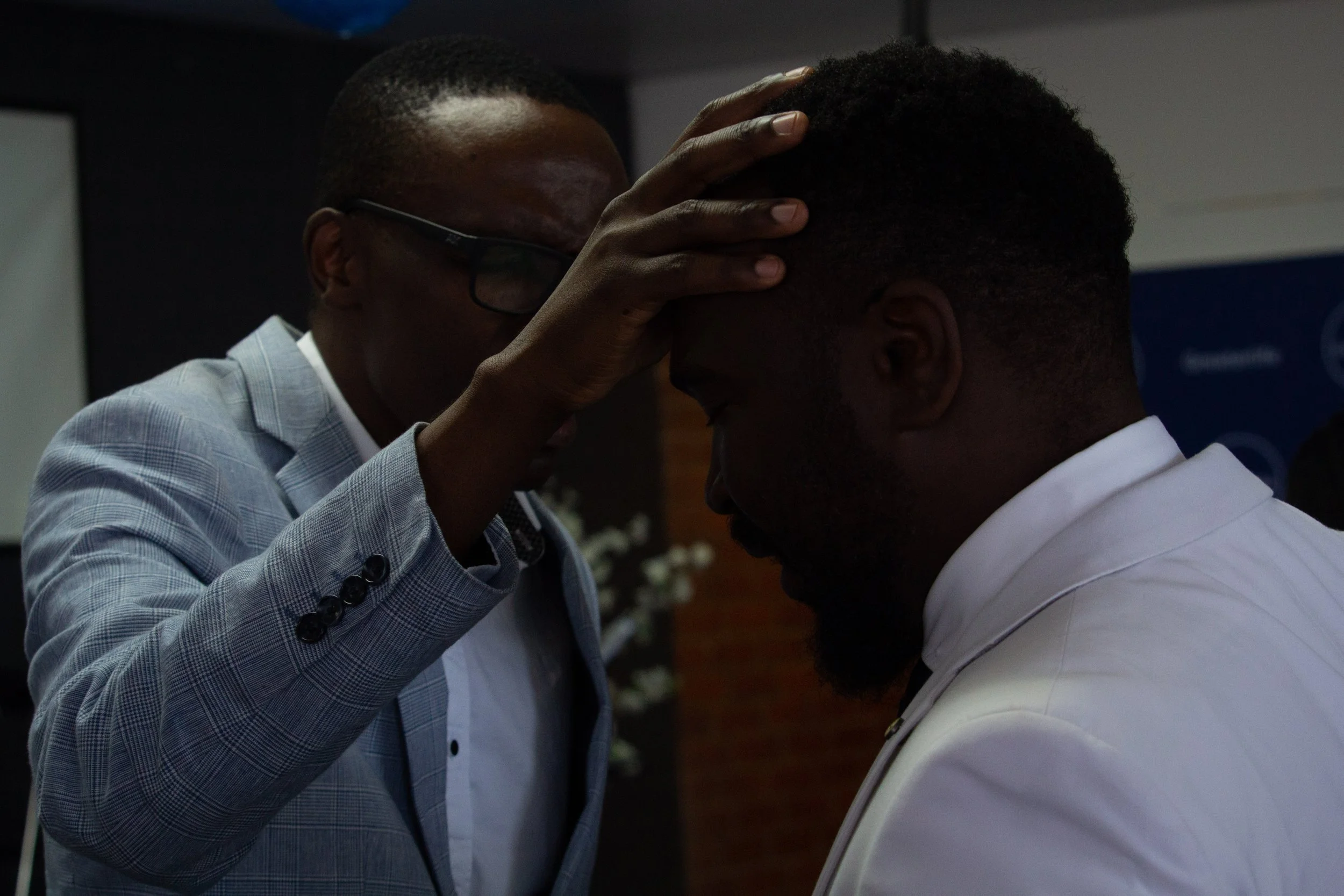 Pastor Simon Phiri placing his hand on another man's head in a moment of prayer or blessing, indoors with a dark background. Pictured at a Greaterlife Church Sunday Service.