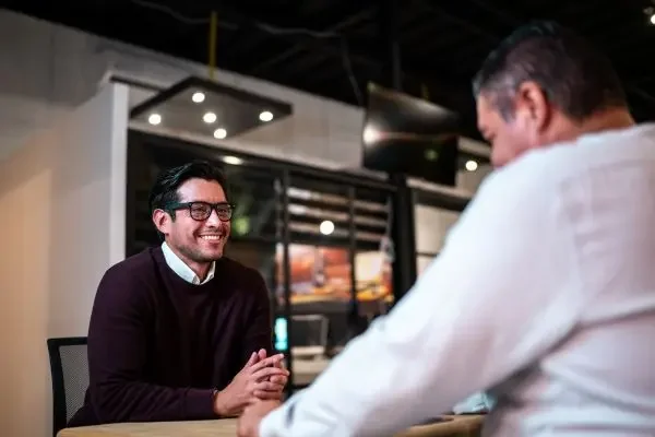 Two men sitting at a table engaged in an informational interview, smiling, in a modern indoor office.