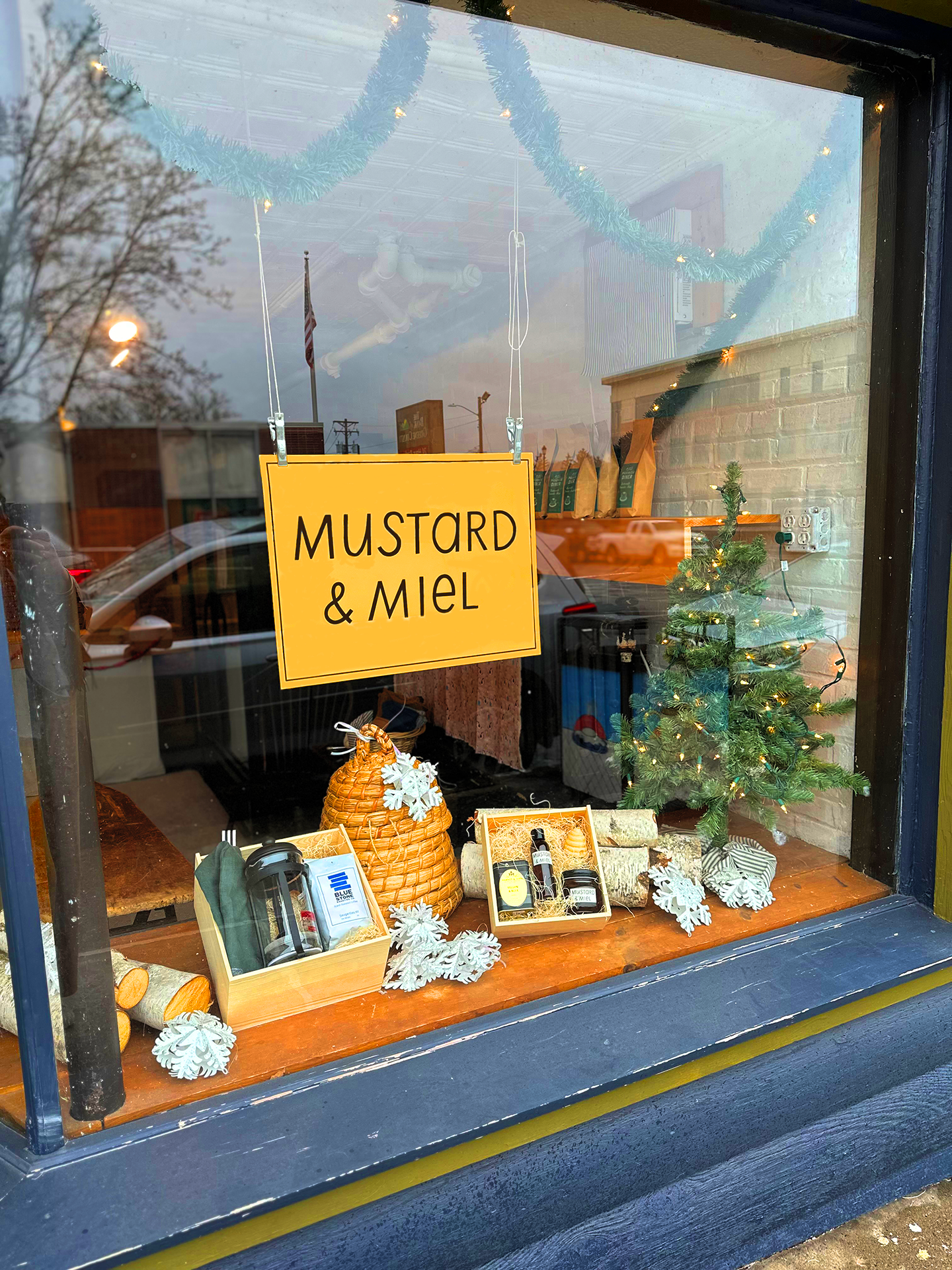 Exterior of the Mustard & Miel storefront with a festive holiday window display of gift boxes, beeswax candles, and birch logs.