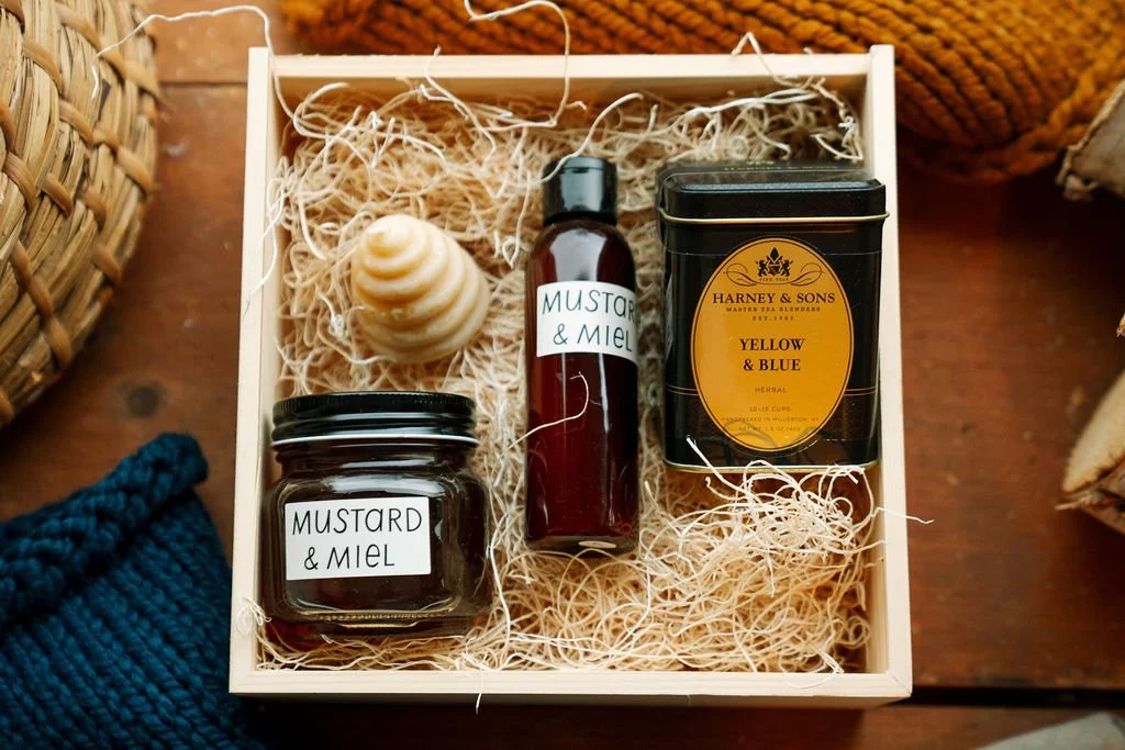 Curated holiday gift box featuring Mustard & Miel honey and body wash, Harney & Sons tea, and a skep-shaped candle nestled in straw.