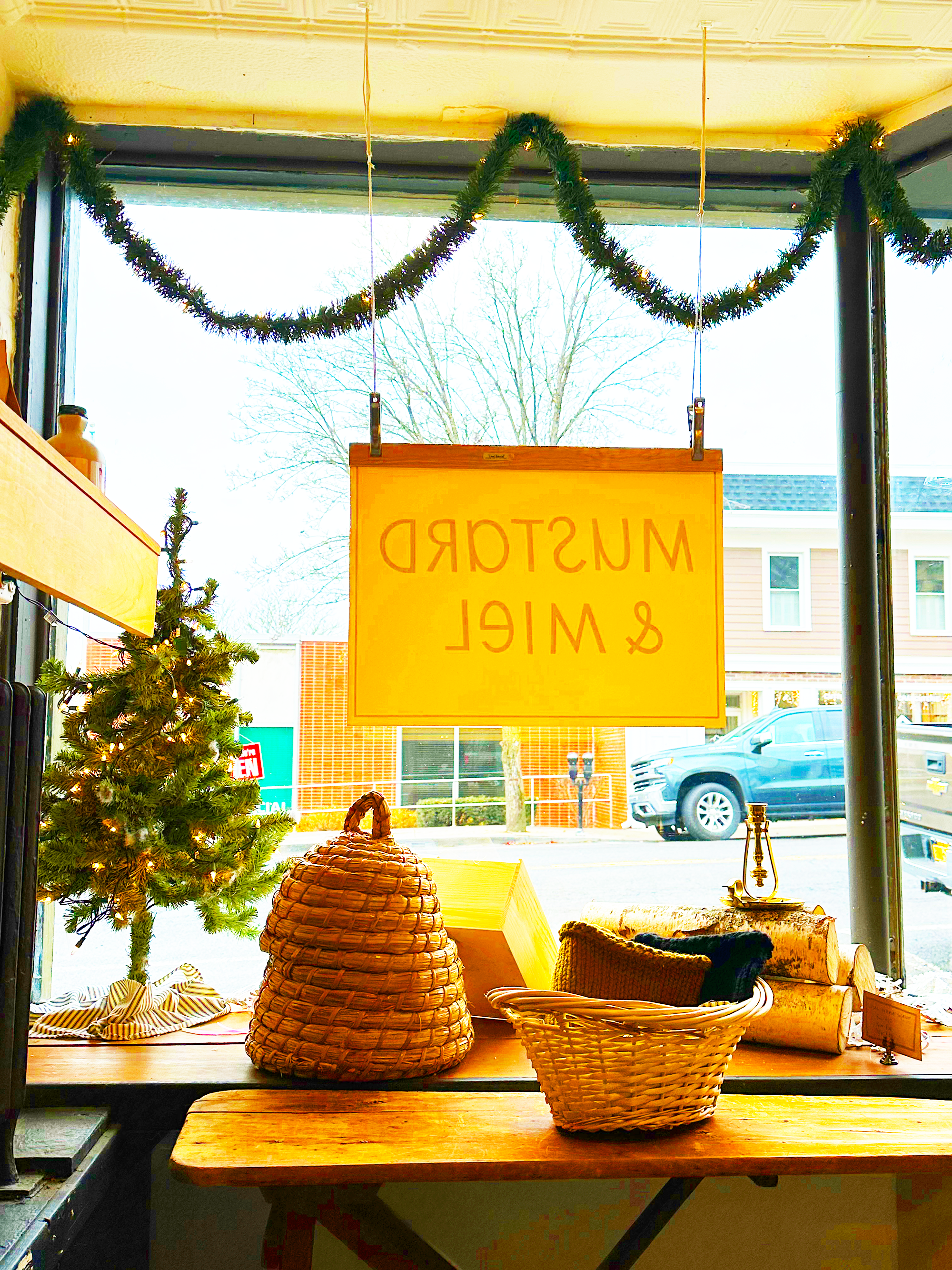 Holiday window display from inside the Mustard & Miel store with beehives, baskets, and garlands framing the downtown Catskill view.