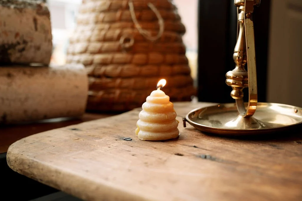 Mustard & Miel's beeswax candle shaped like a skep beehive, lit, on a rustic wooden table, beside a brass candlestick holder.