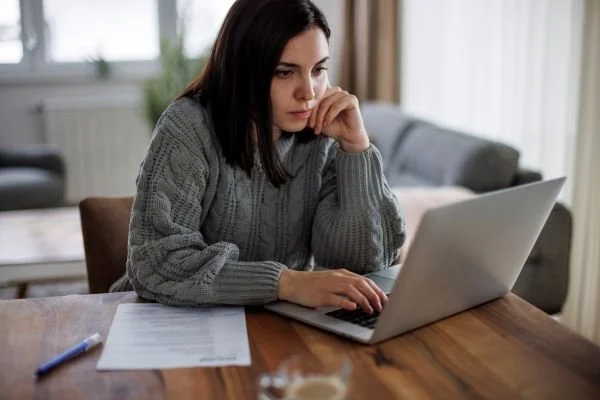 Woman in a gray sweater working on a laptop at a wooden table with papers and a pen, in a room with a window and curtains, symbolizing a job seeker looking for a career in higher education.