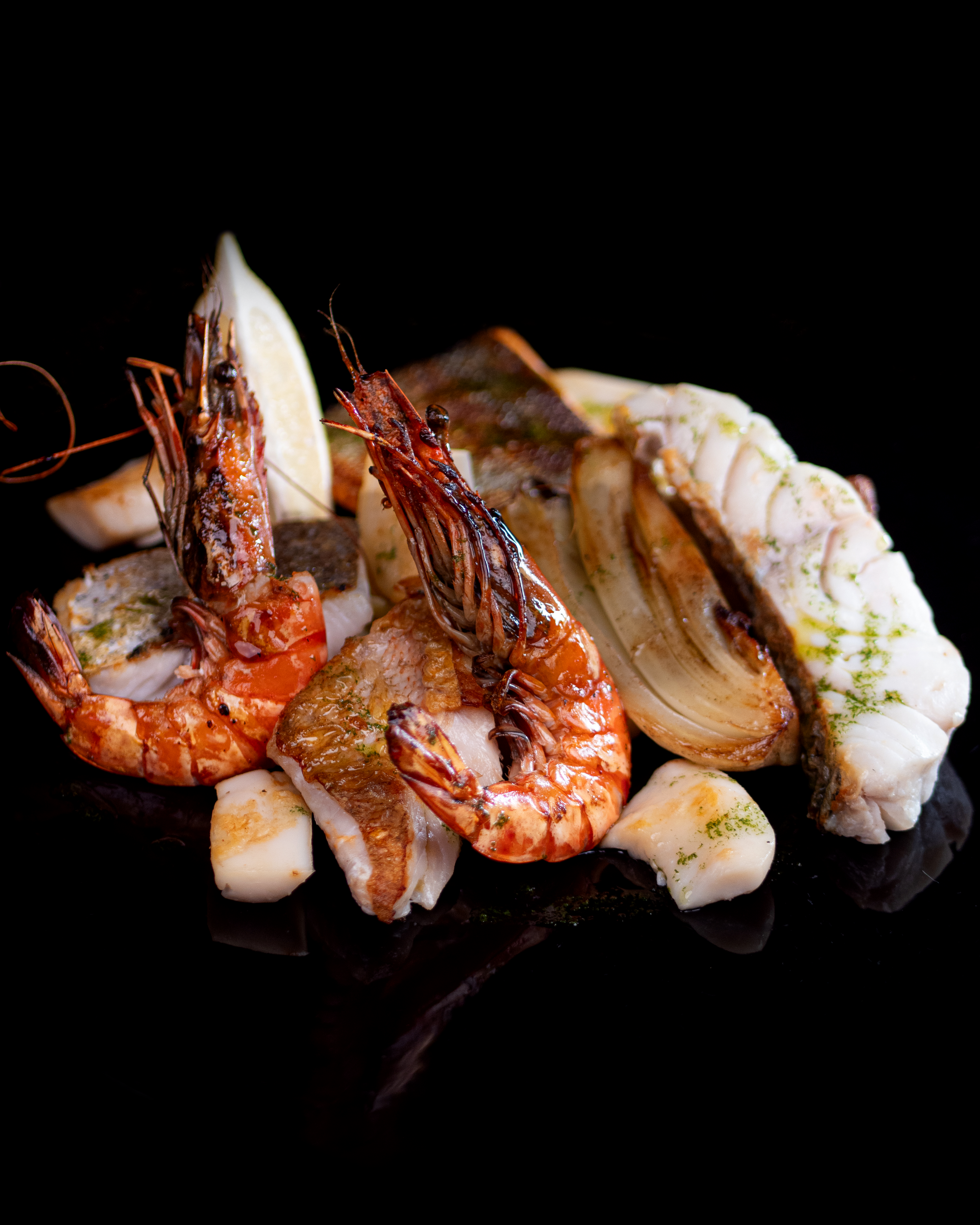 Gambas ou crevettes grillées avec du citron, accompagnées de morceaux de poisson ou de calamars, servi sur une surface noire.