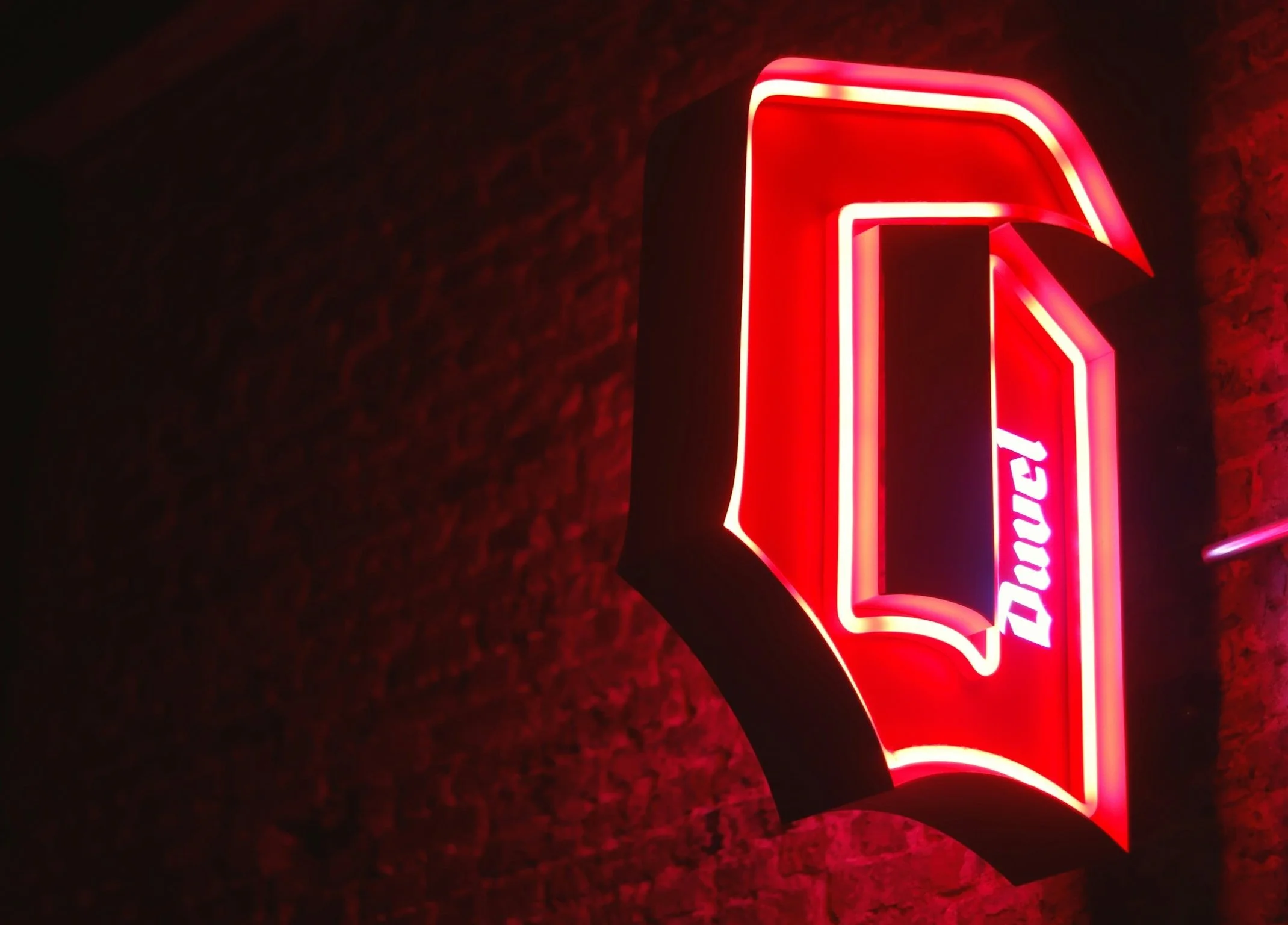 Red illuminated sign of DUVEL, mounted on a dark brick wall.