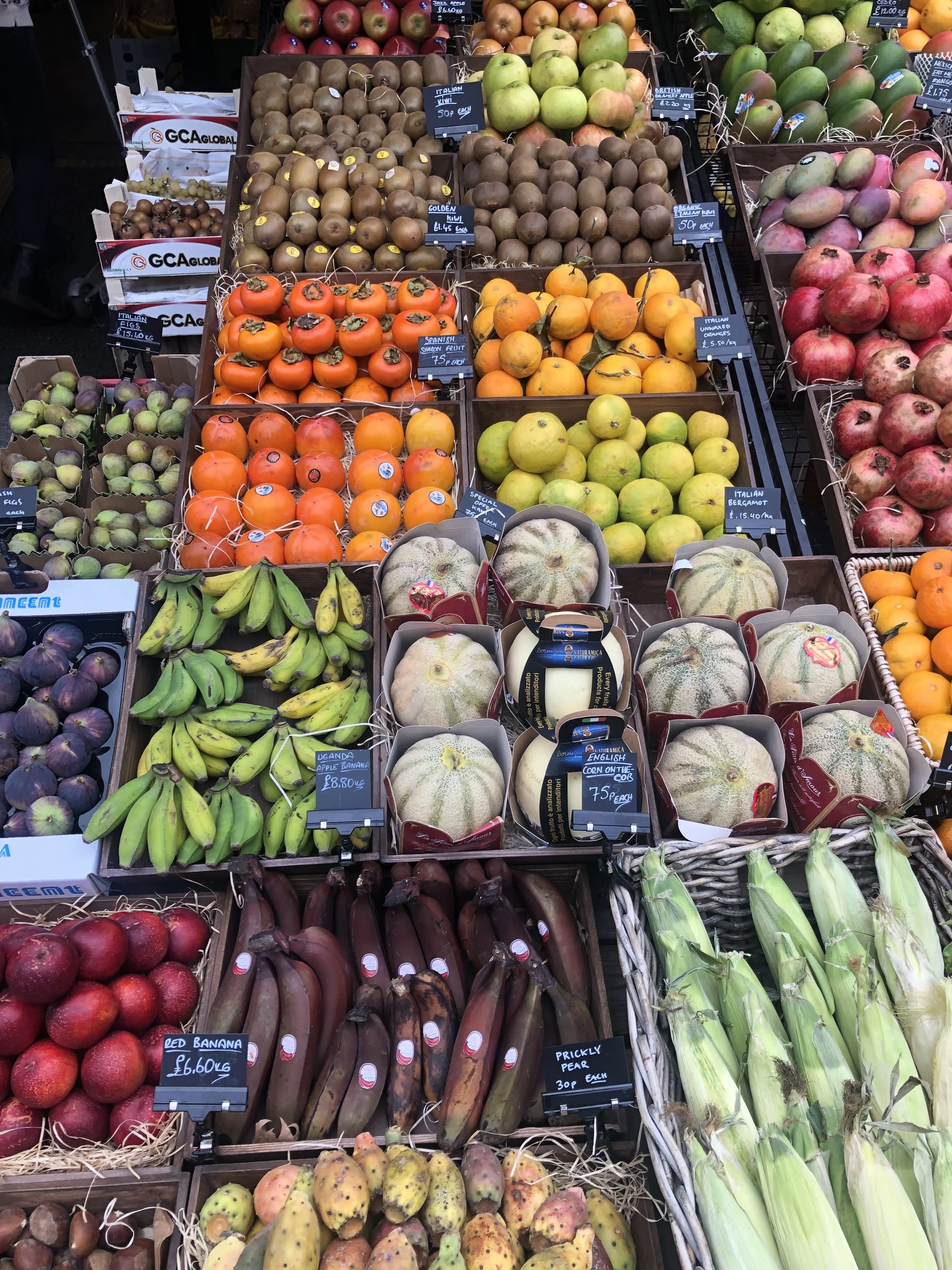 A display of various fruits at a market, including apples, kiwis, oranges, bananas, watermelons, eggplants, shallots, and corn, arranged in wooden and cardboard baskets.