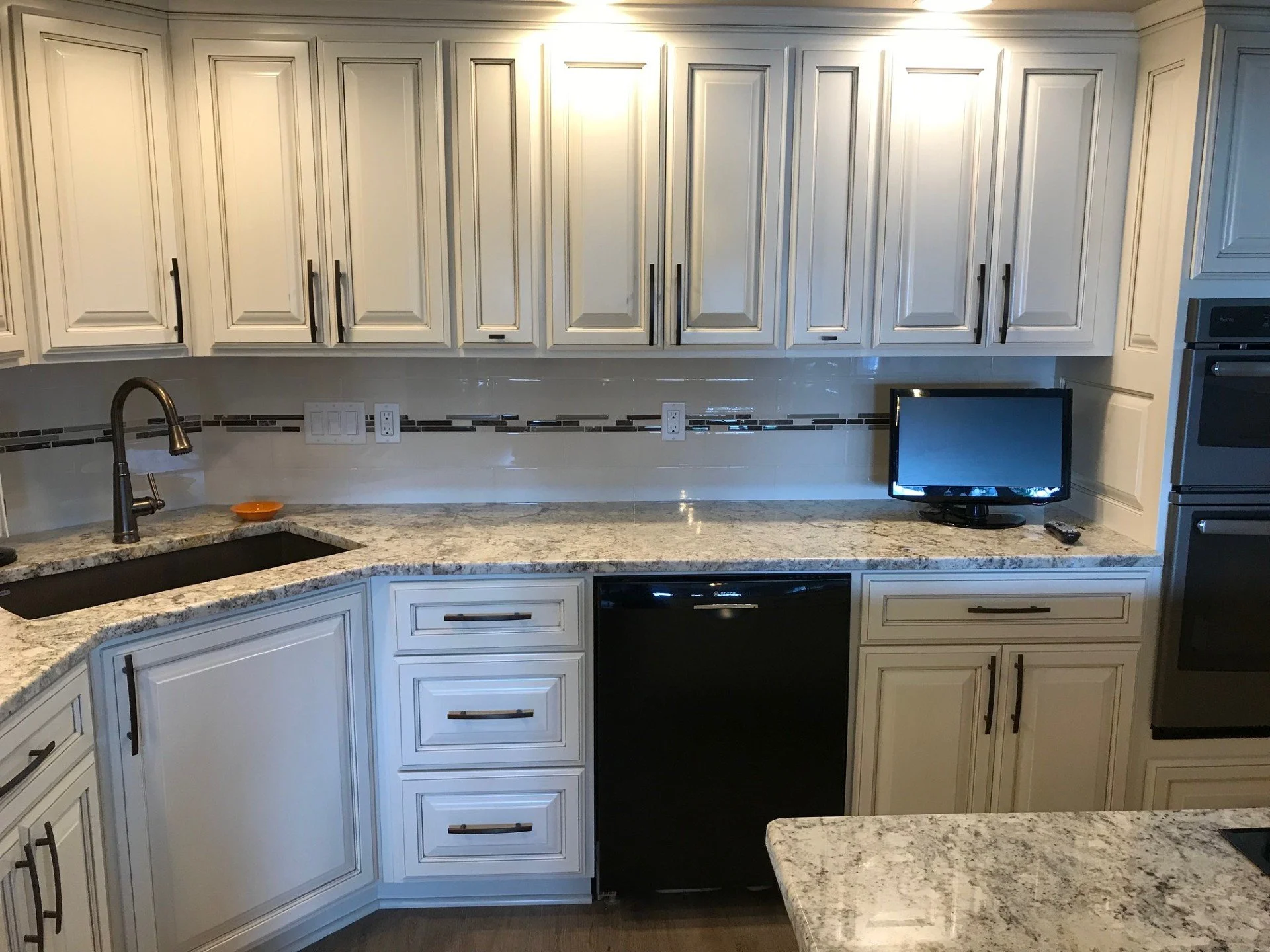 Kitchen with white cabinets, granite countertops, and a small TV on the counter. There is a black dishwasher below the counter and a double oven built into the cabinetry on the right.