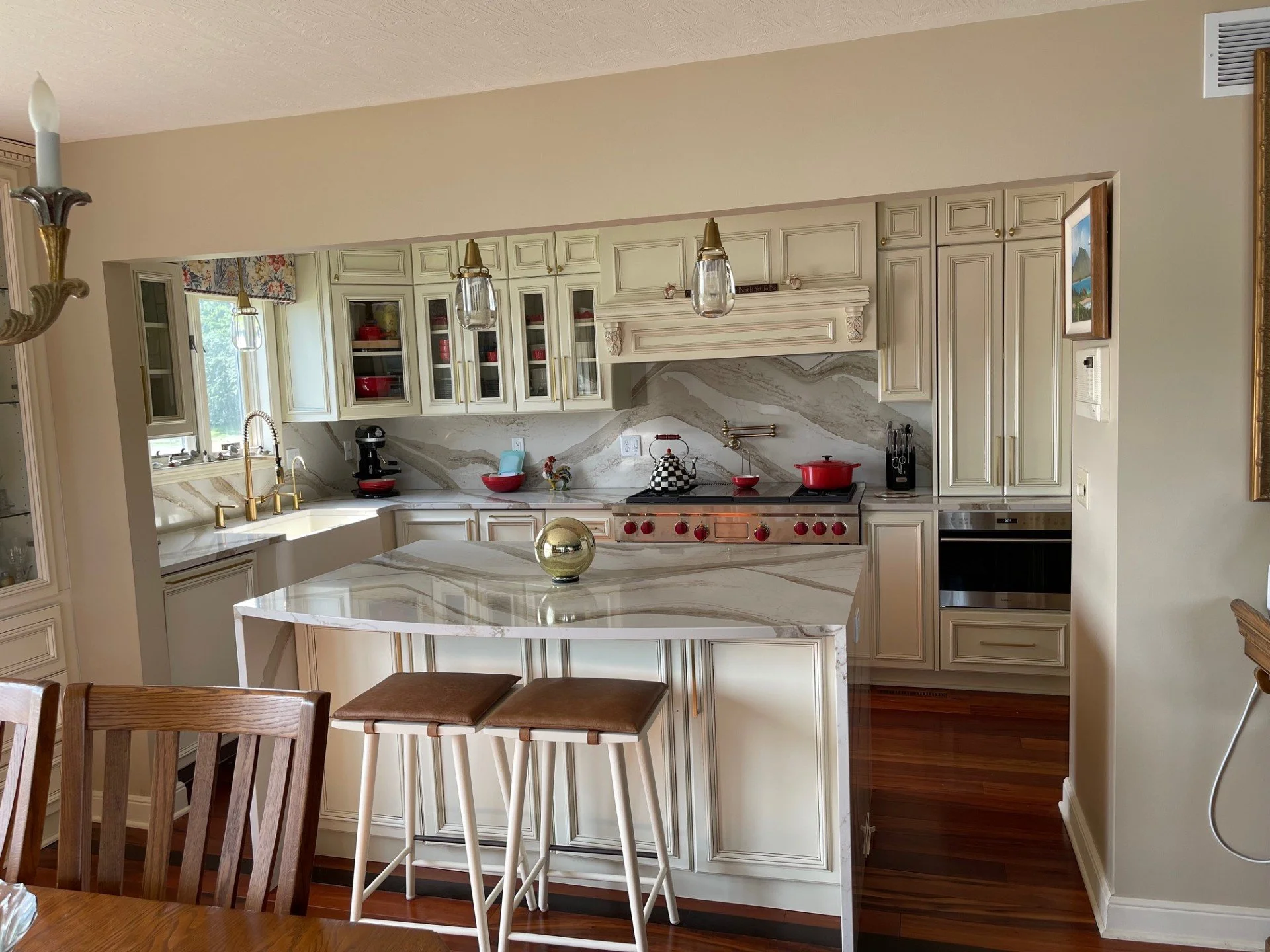 A kitchen with cream-colored cabinets, a marble countertop island, and red accents. There are three bar stools at the island, a double sink beneath a window, and a stove with red knobs. Pendant lights hang above the island, and various kitchen utensi