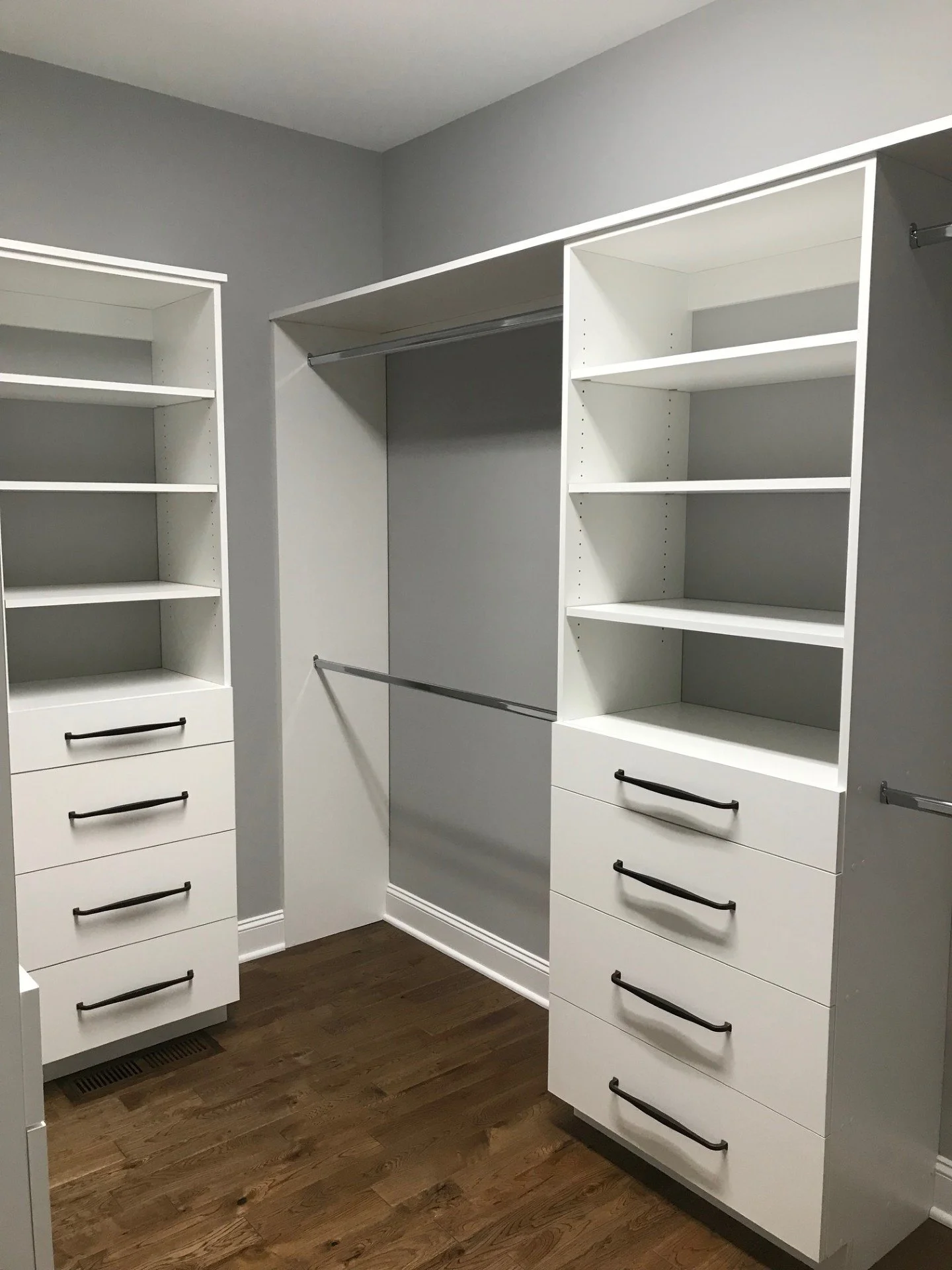 Empty walk-in closet with white shelving units, drawers, and hanging rods against gray walls and a wooden floor.