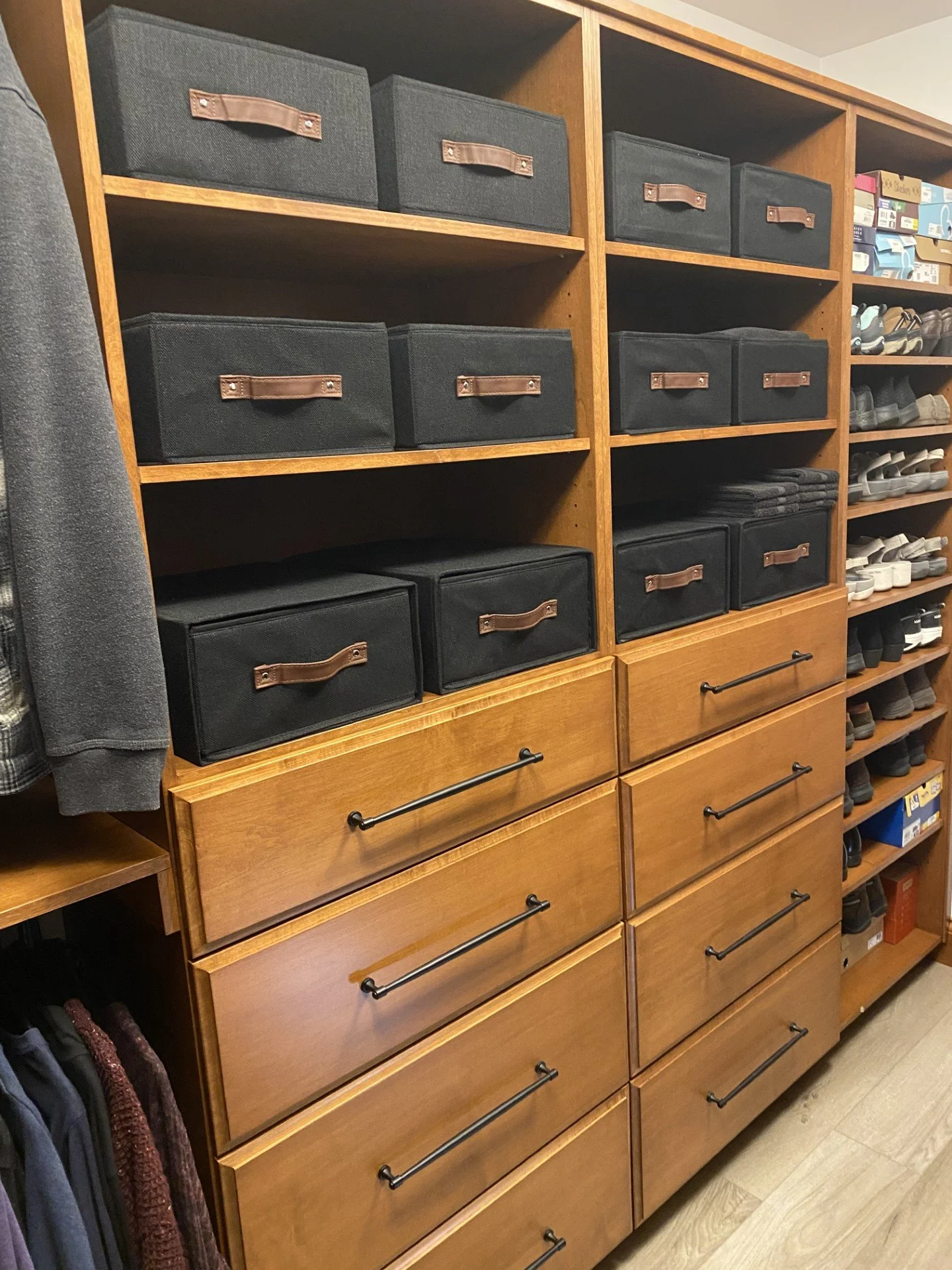 Wooden shelving unit with black storage boxes on top and drawers with black handles below, next to shoe shelves filled with various sneakers.