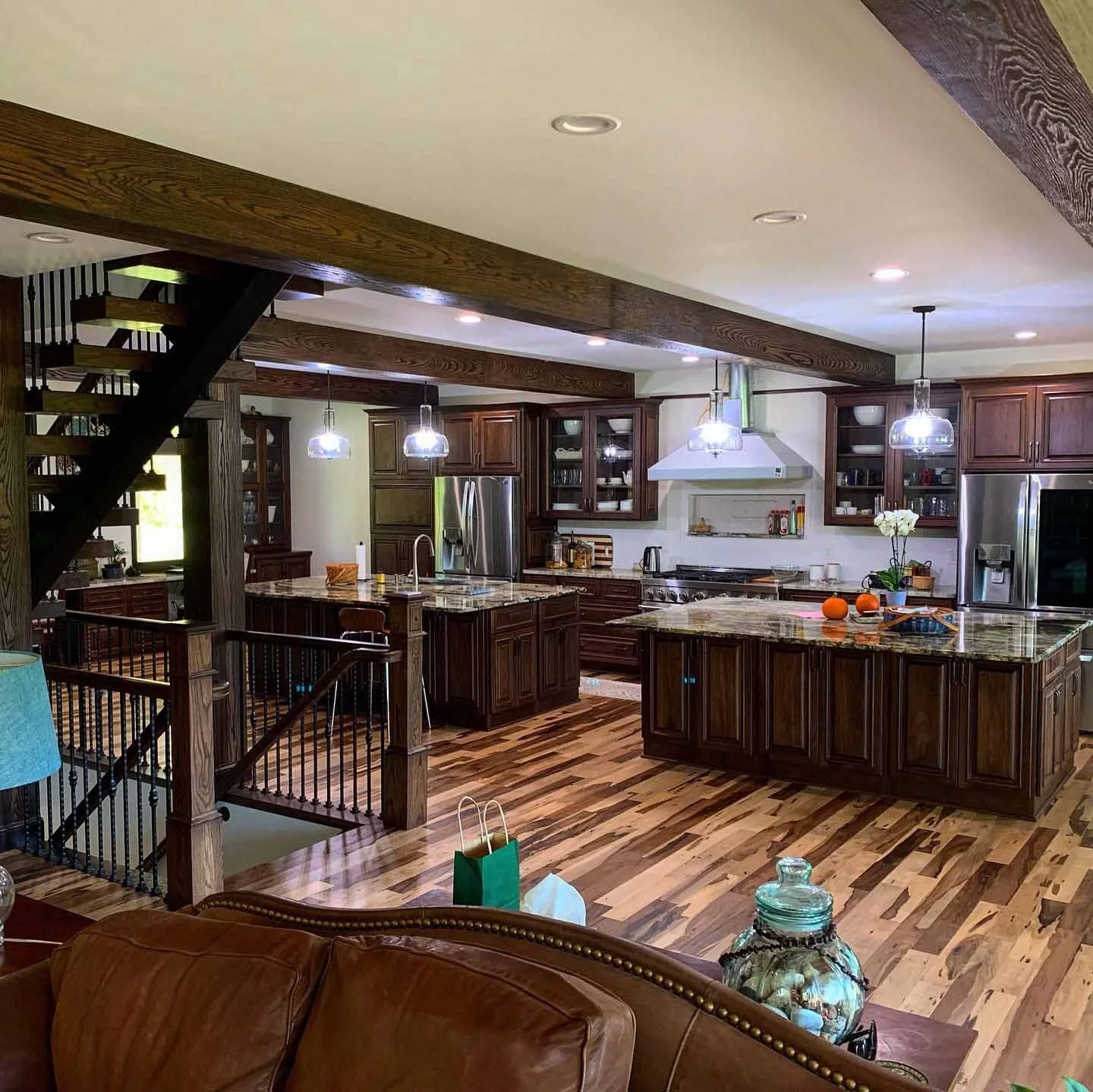 A spacious kitchen with dark wooden cabinets, a large island with granite countertop, stainless steel appliances, pendant lights, and hardwood flooring. There are decorative items and a small gift bag on the island and a leather couch partially visible in the foreground.