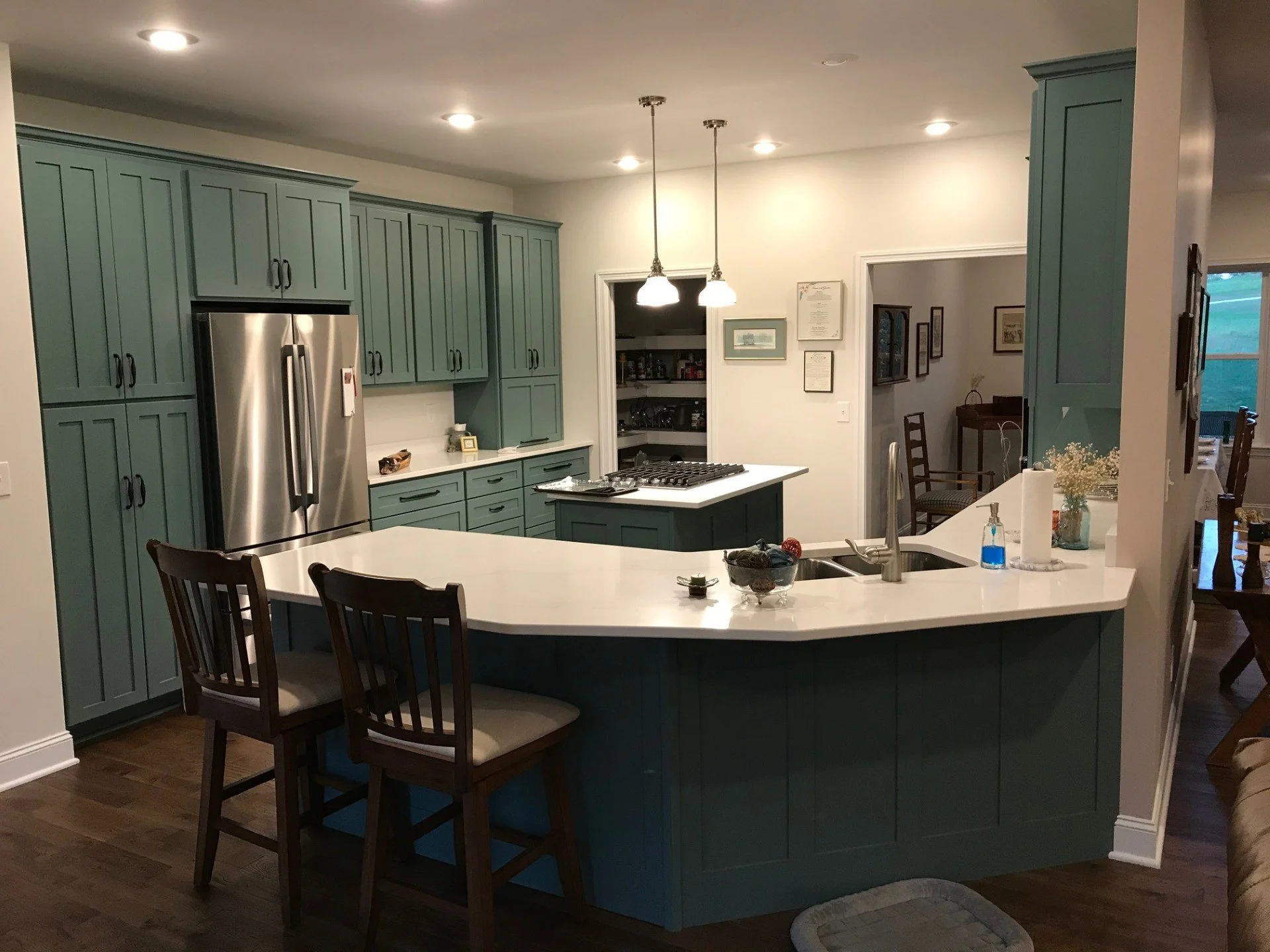Modern kitchen with green cabinetry, white countertops, stainless steel refrigerator, island with two chairs, and hanging pendant lights.