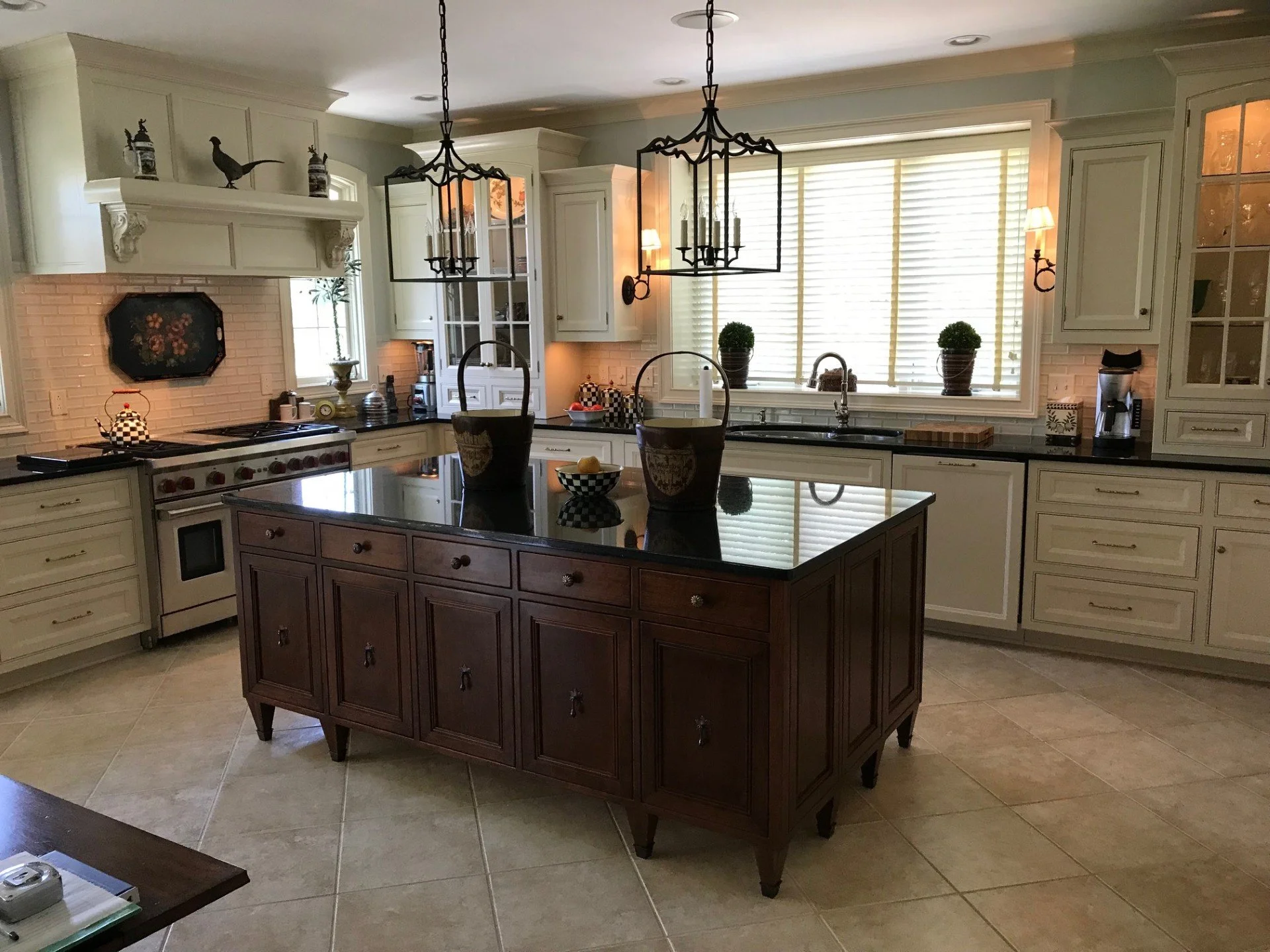 A spacious kitchen with white cabinets, black countertops, and a central dark wood island. Two black wrought iron chandeliers hang above the island, which has decorative potted plants and jars. Large window with blinds allows natural light, with addi
