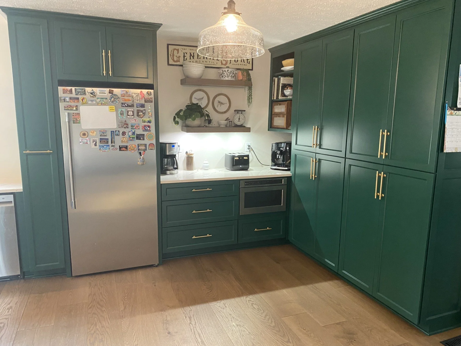 Green kitchen with a stainless steel refrigerator covered in various magnets, a coffee maker, a microwave, and open shelving with plants, books, and decorative items. Hardwood floor and a light fixture hang from the ceiling.
