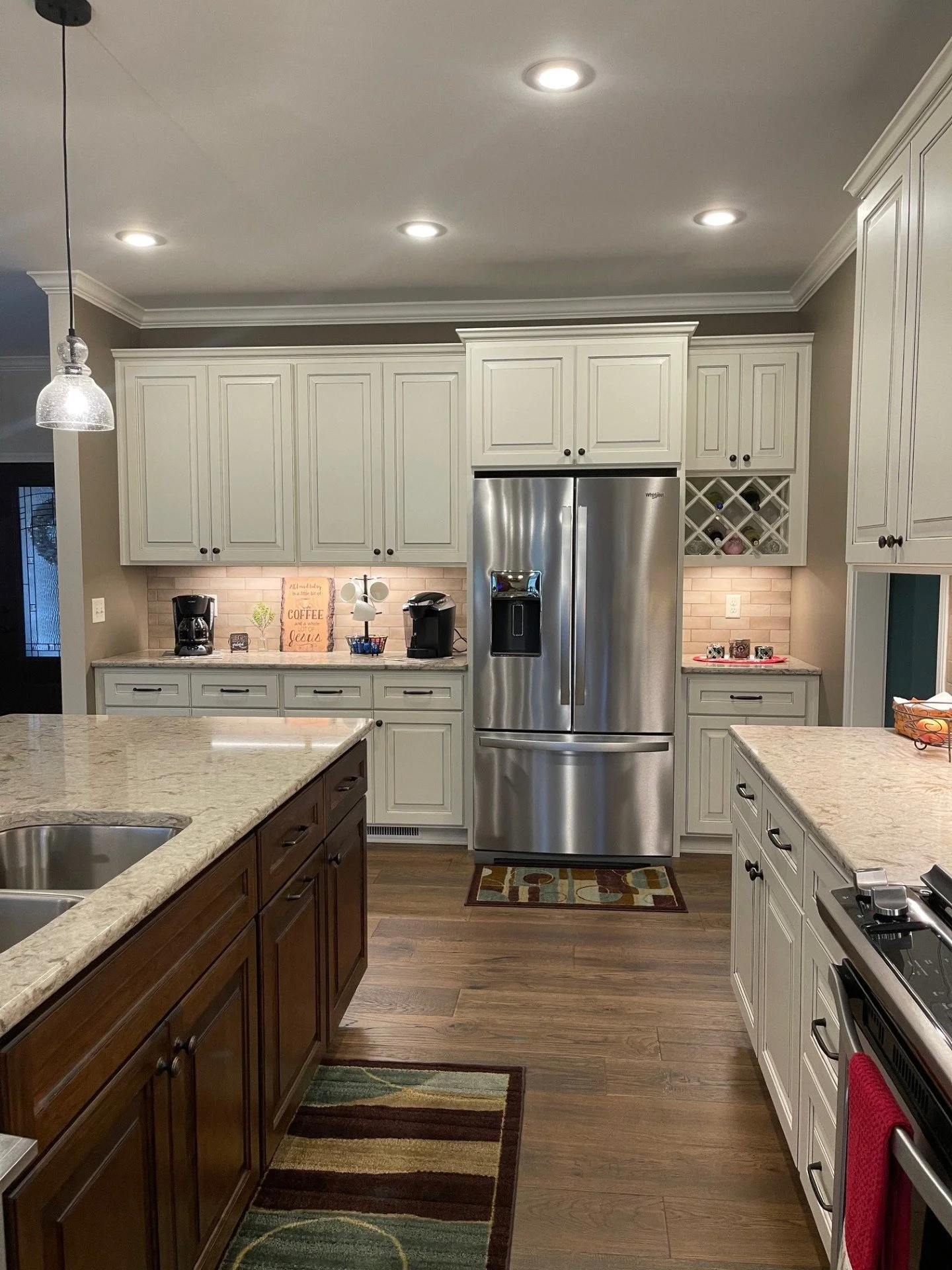 A kitchen with a mix of dark wood and white cabinets, granite countertops, and stainless steel appliances including a refrigerator and an oven. There are two rugs on the wooden floor.