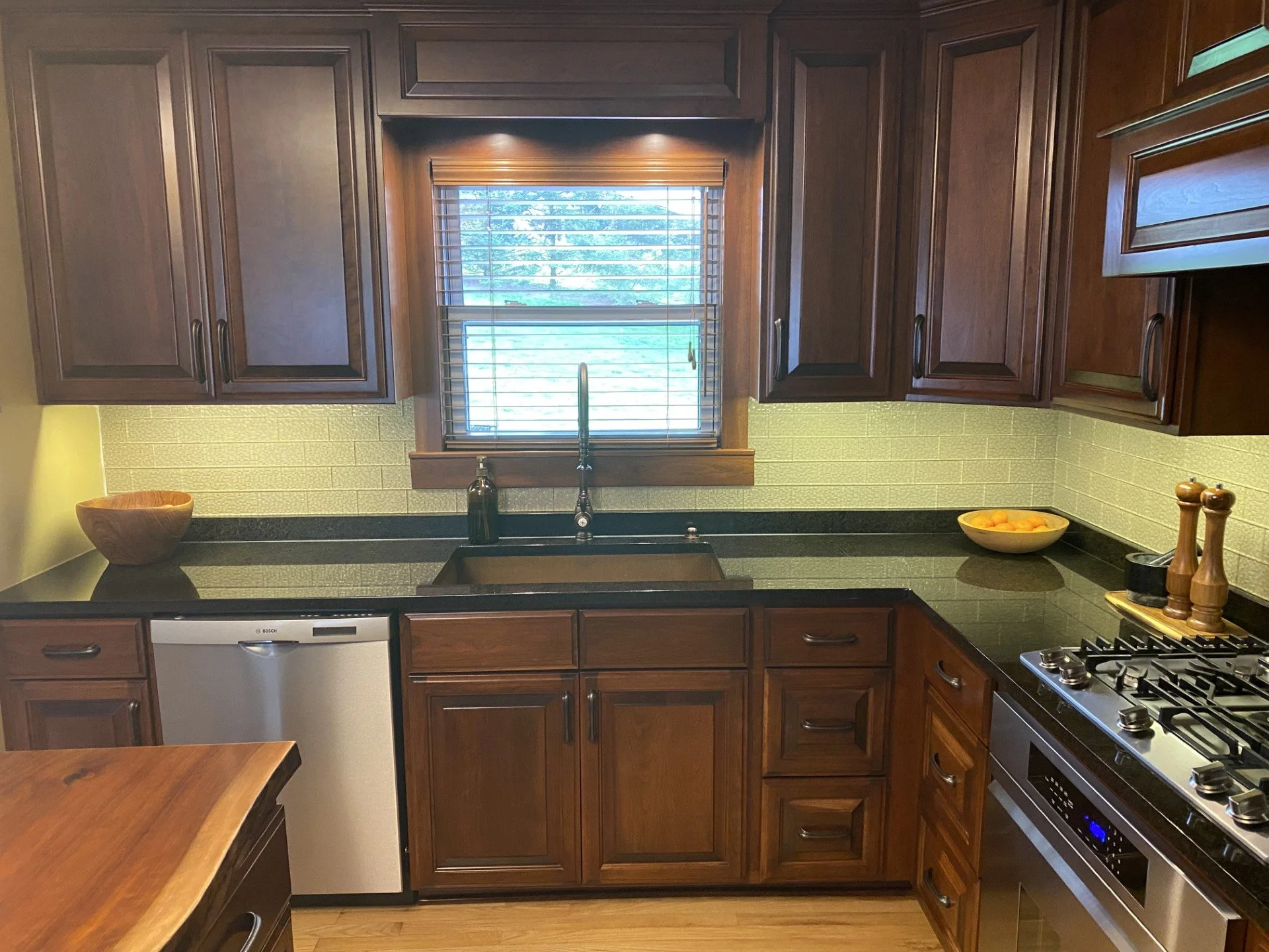 Kitchen with dark wood cabinets, black countertops, a window with blinds above the sink, a small dishwasher, a stove, and a bowl of orange fruit.
