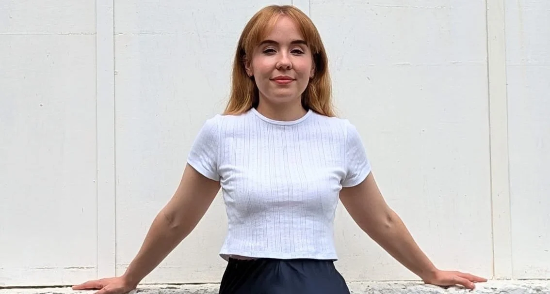 Young woman with red hair wearing a white top and black skirt standing against a white background.