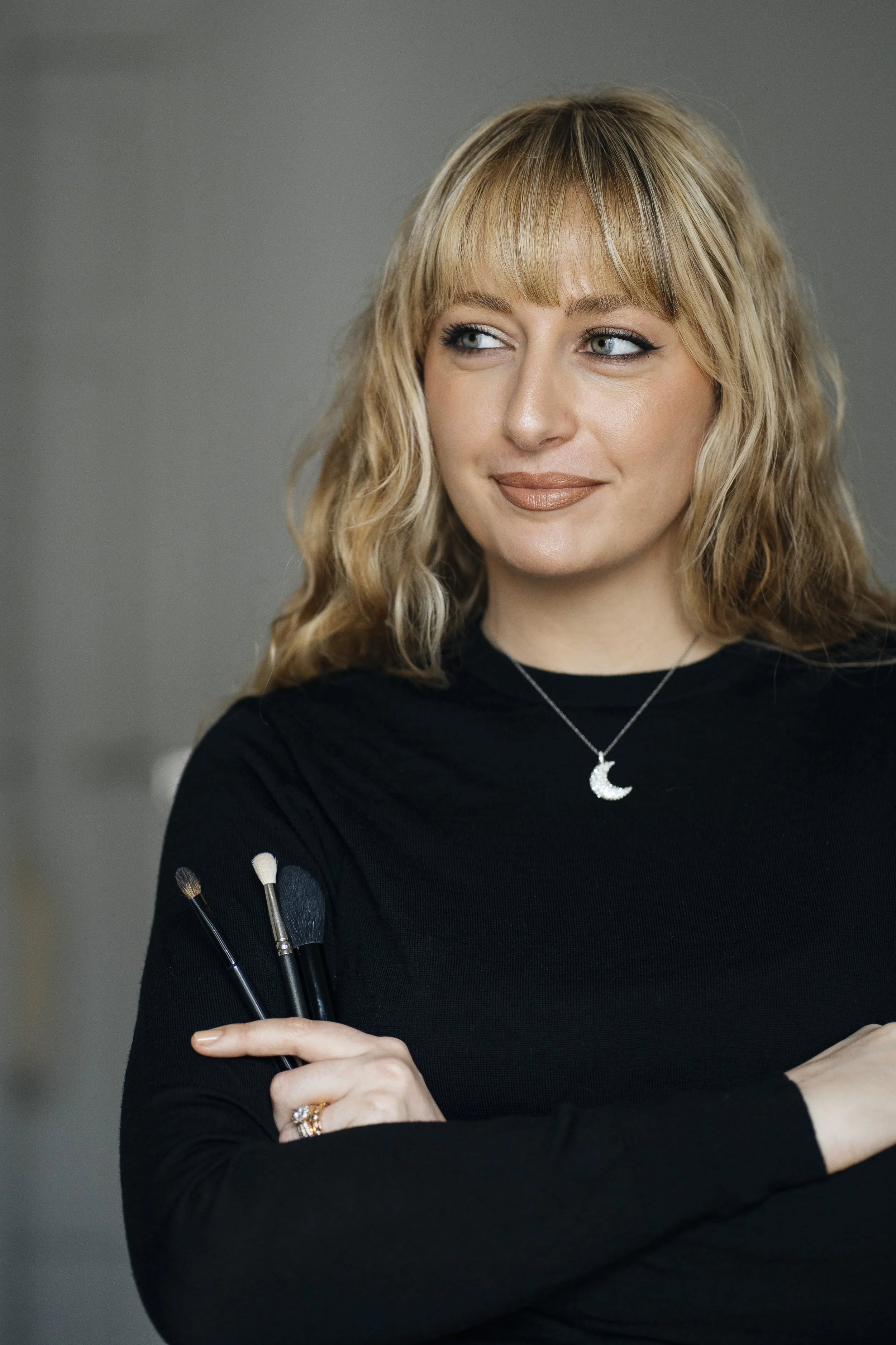 Une femme blonde portant un pendentif en forme de lune, habillée en noir, tenant des pinceaux à maquillage, avec un sourire confiant et regardant sur le côté.