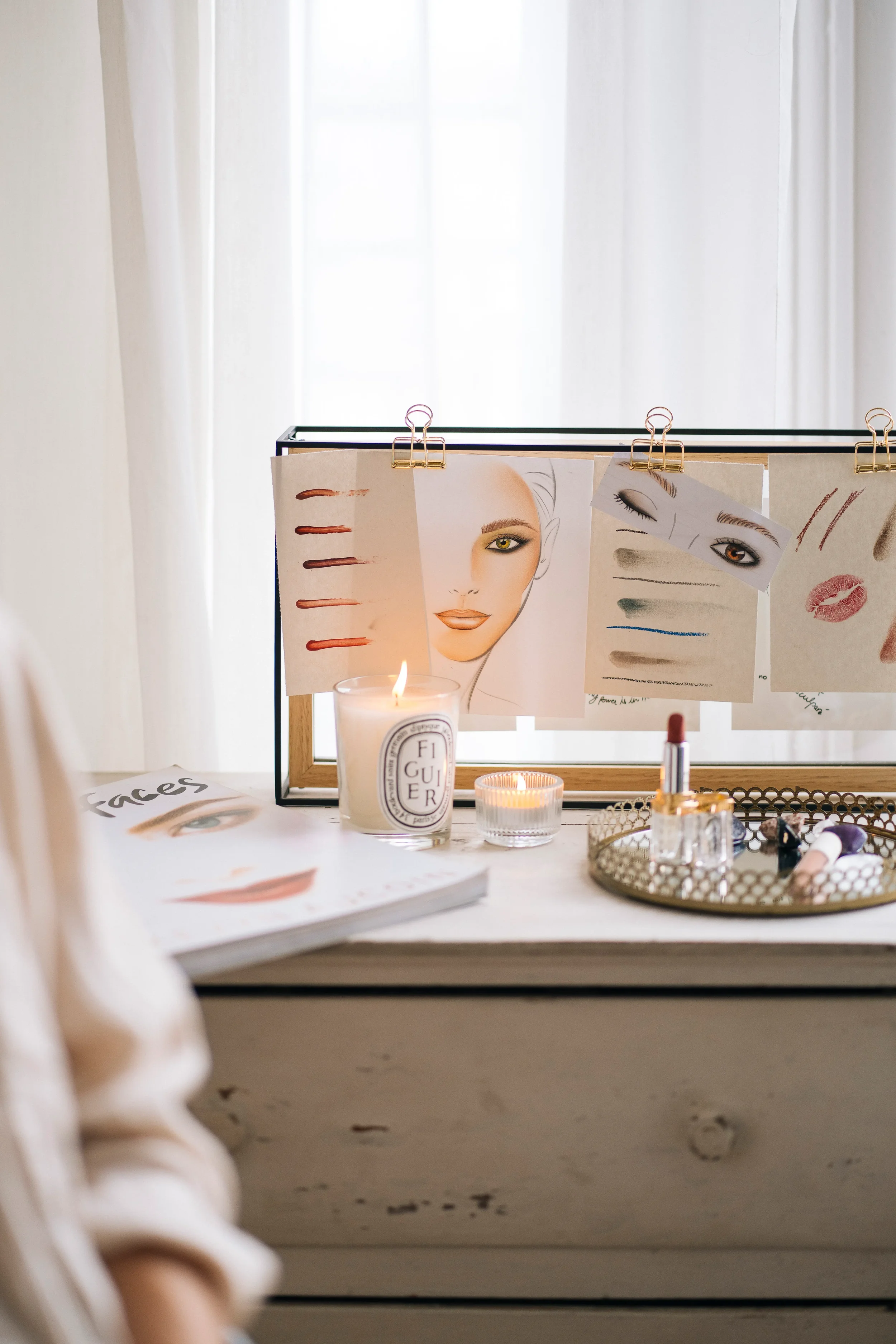 Table avec magazines et croquis de maquillage, bougies allumées, et un miroir avec des échantillons de maquillage suspendus.