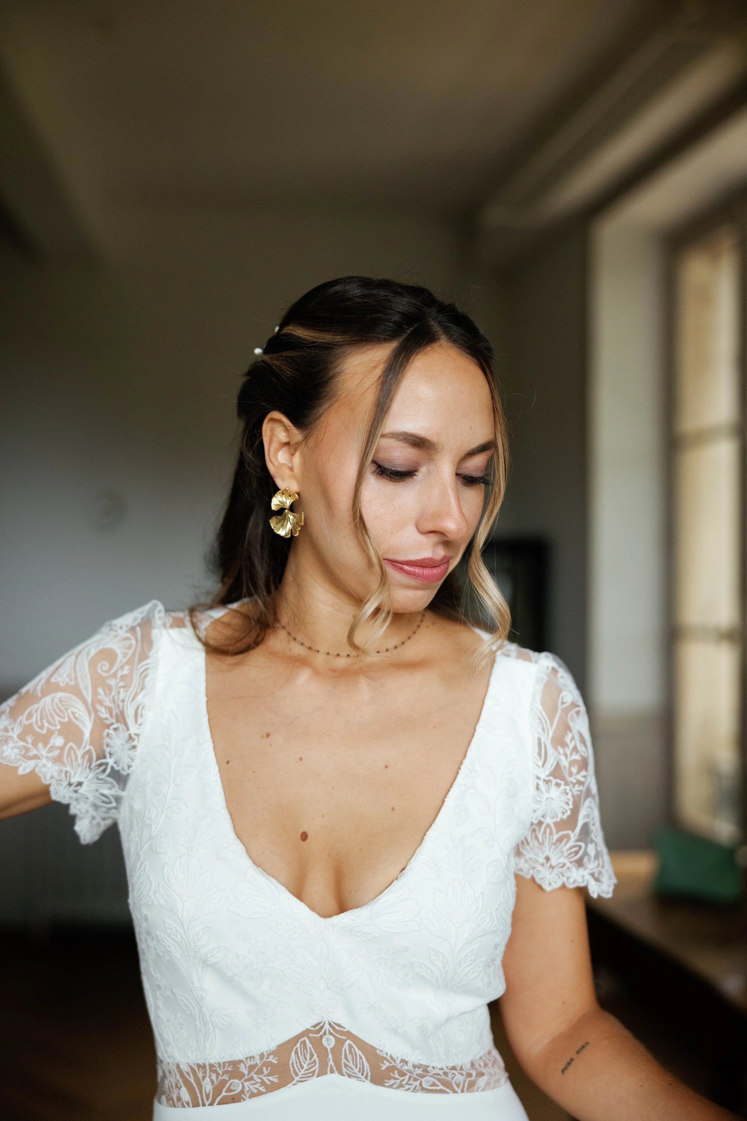 Jeune femme portant une robe blanche en dentelle et des boucles d'oreilles dorées, debout près d'une fenêtre, regardant vers le bas.