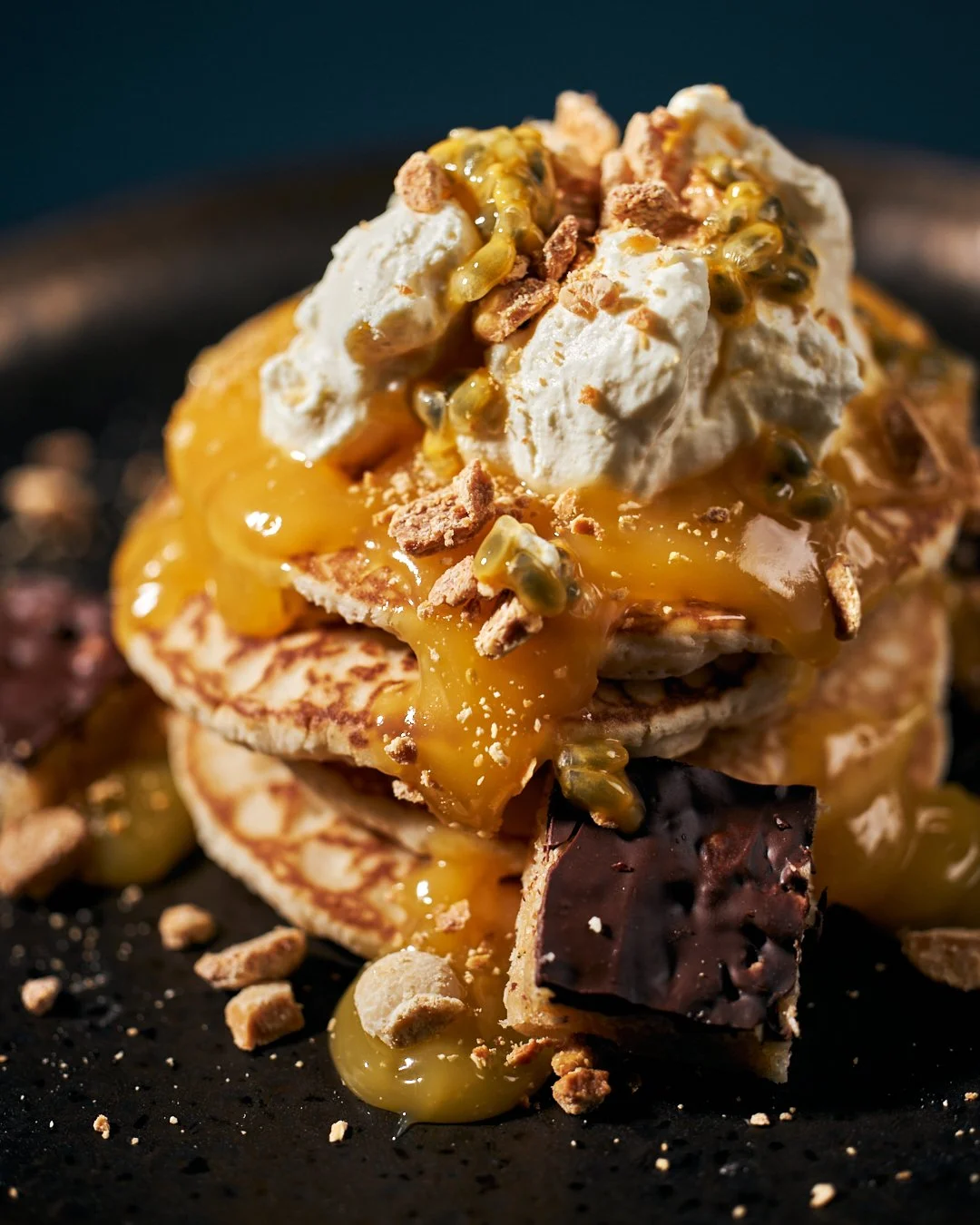 Stack of pancakes topped with whipped cream, passion fruit, caramel sauce, chopped nuts, and a piece of chocolate