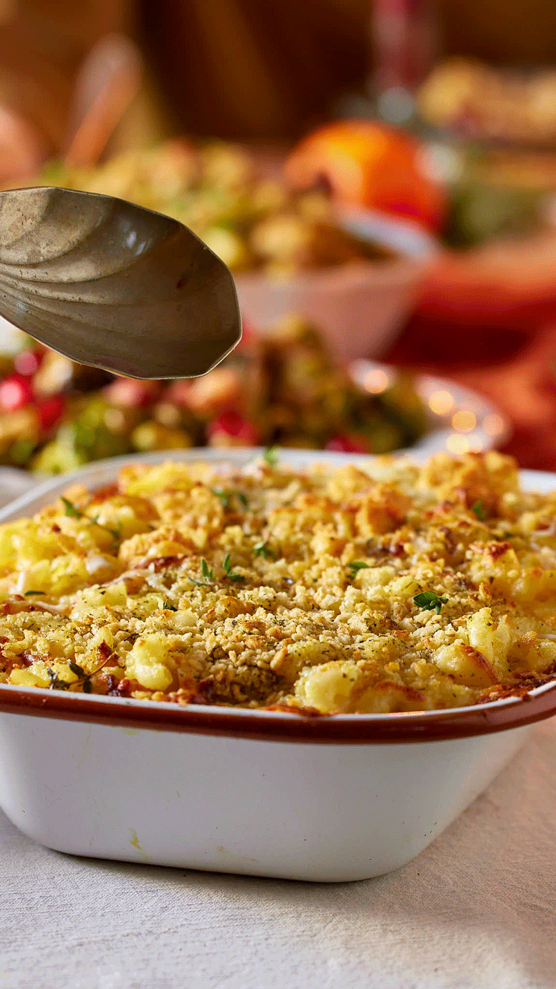 A casserole dish filled with a baked potato topping mixed with cheese, bacon, and herbs, being served with a spoon.
