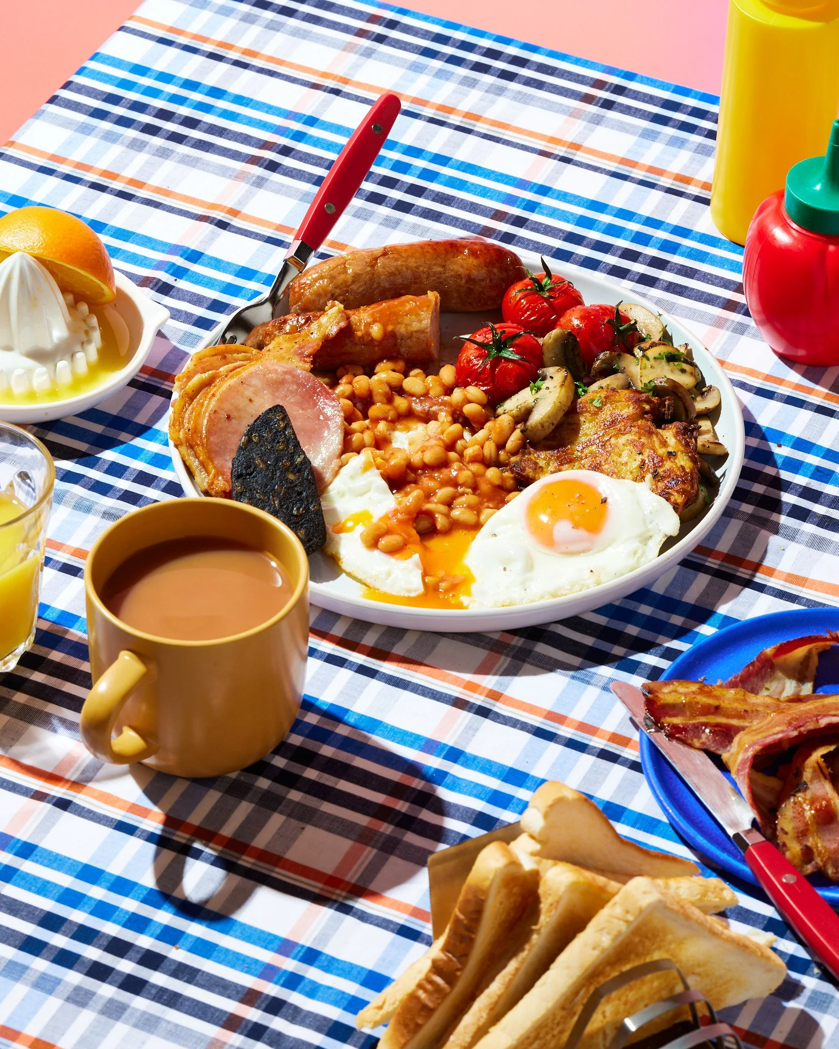 Plate of breakfast foods on a checkered tablecloth, including sunny side up eggs, baked beans, grilled tomatoes, mushrooms, sausage, bacon, and bread slices.