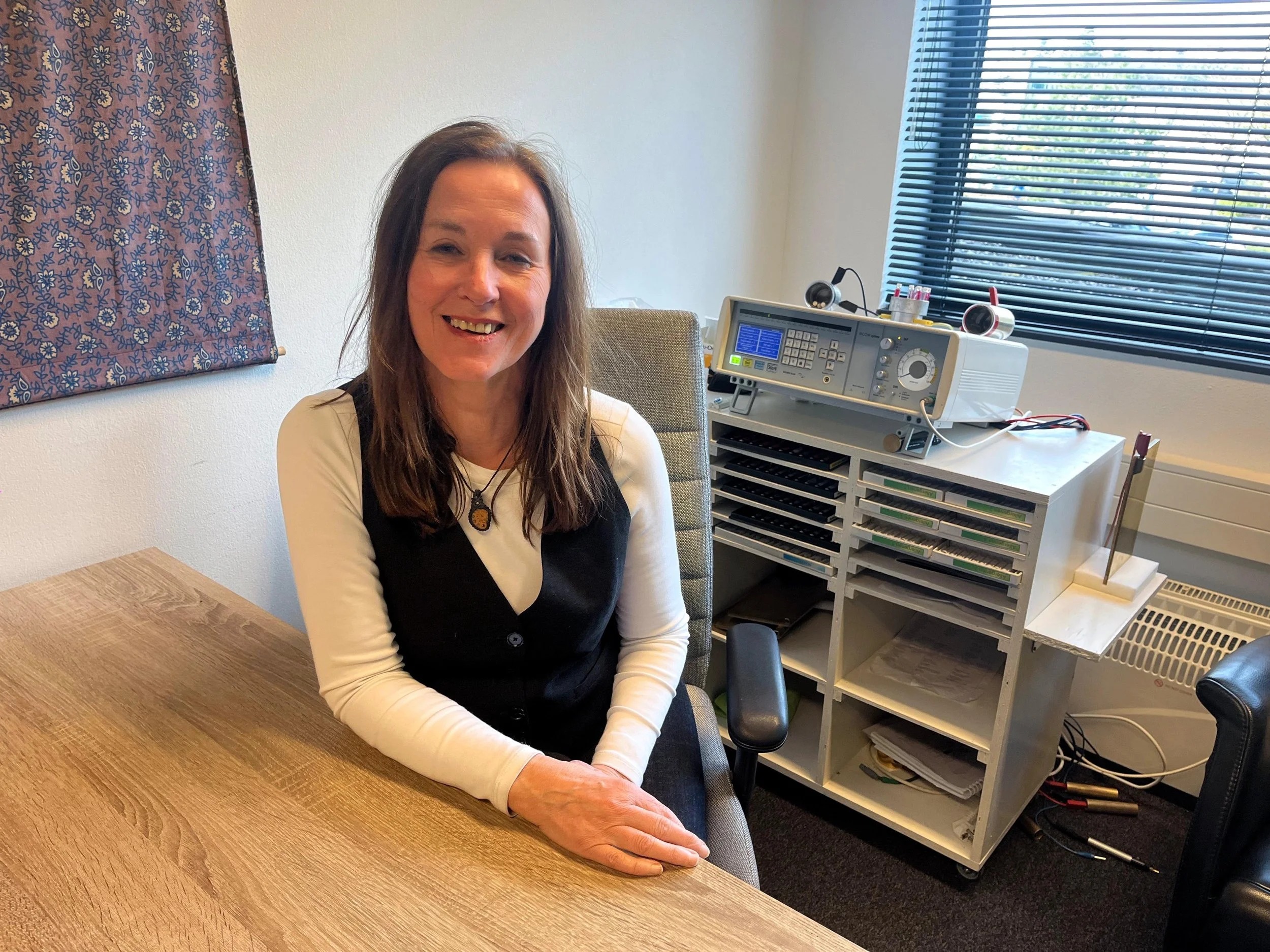 Therapeut Tineke Jacobs zit lachend aan een houten tafel in haar kantoor, met een bioresonantie apparaat en een raam op de achtergrond.