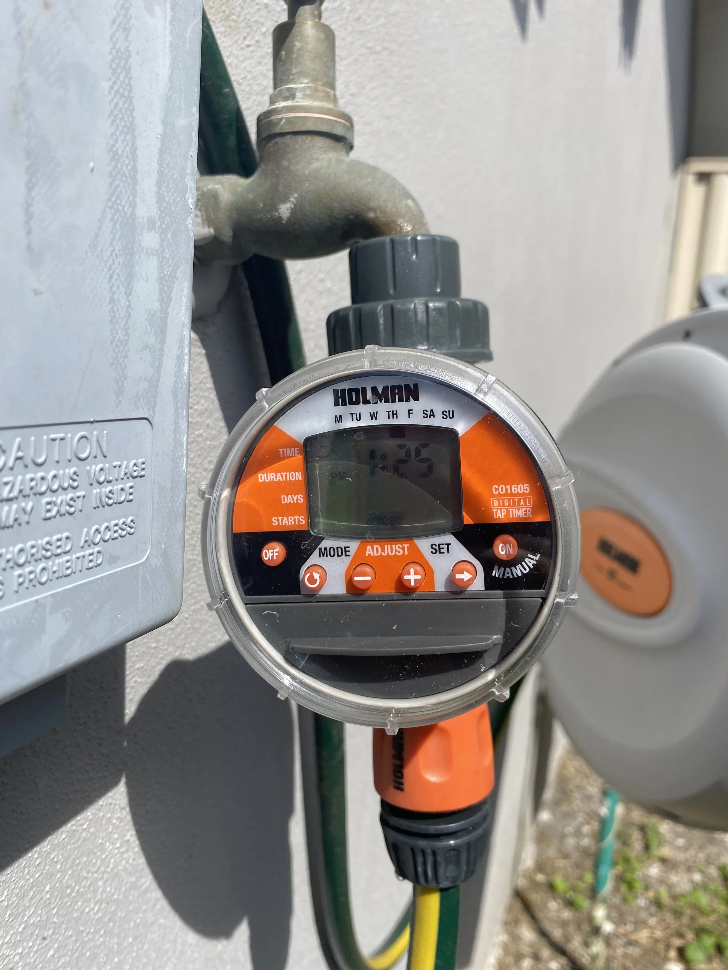 Close-up of an outdoor electrical box with a digital timer showing the time 1:25. A green and yellow cable and an orange cable are attached, with a gardening tool slightly visible in the background.