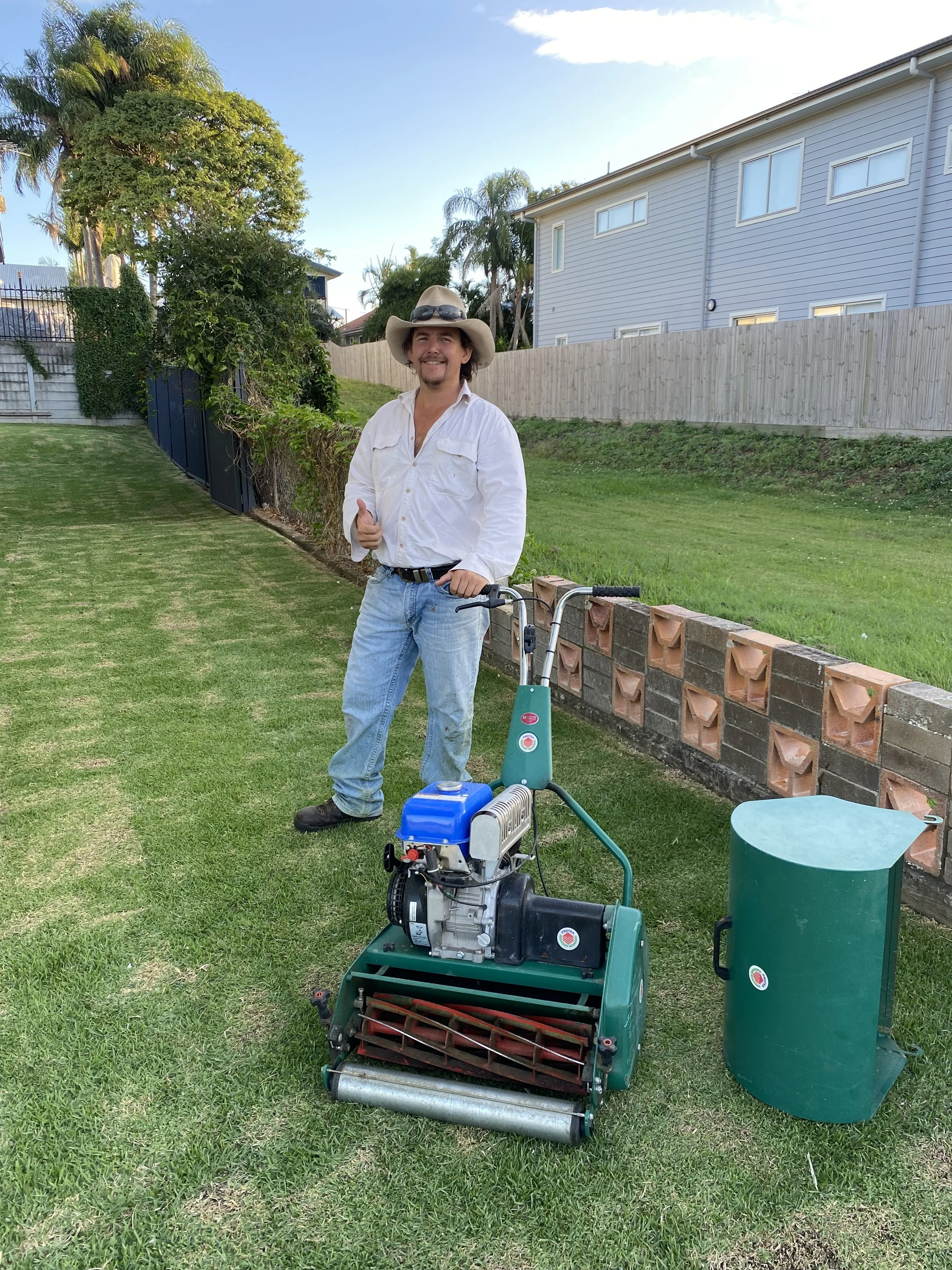 A man wearing a cowboy hat and white shirt giving a thumbs up while standing next to a turf aerator on a grassy yard, with a green trash bin nearby, trees, a wooden fence, and a house in the background.