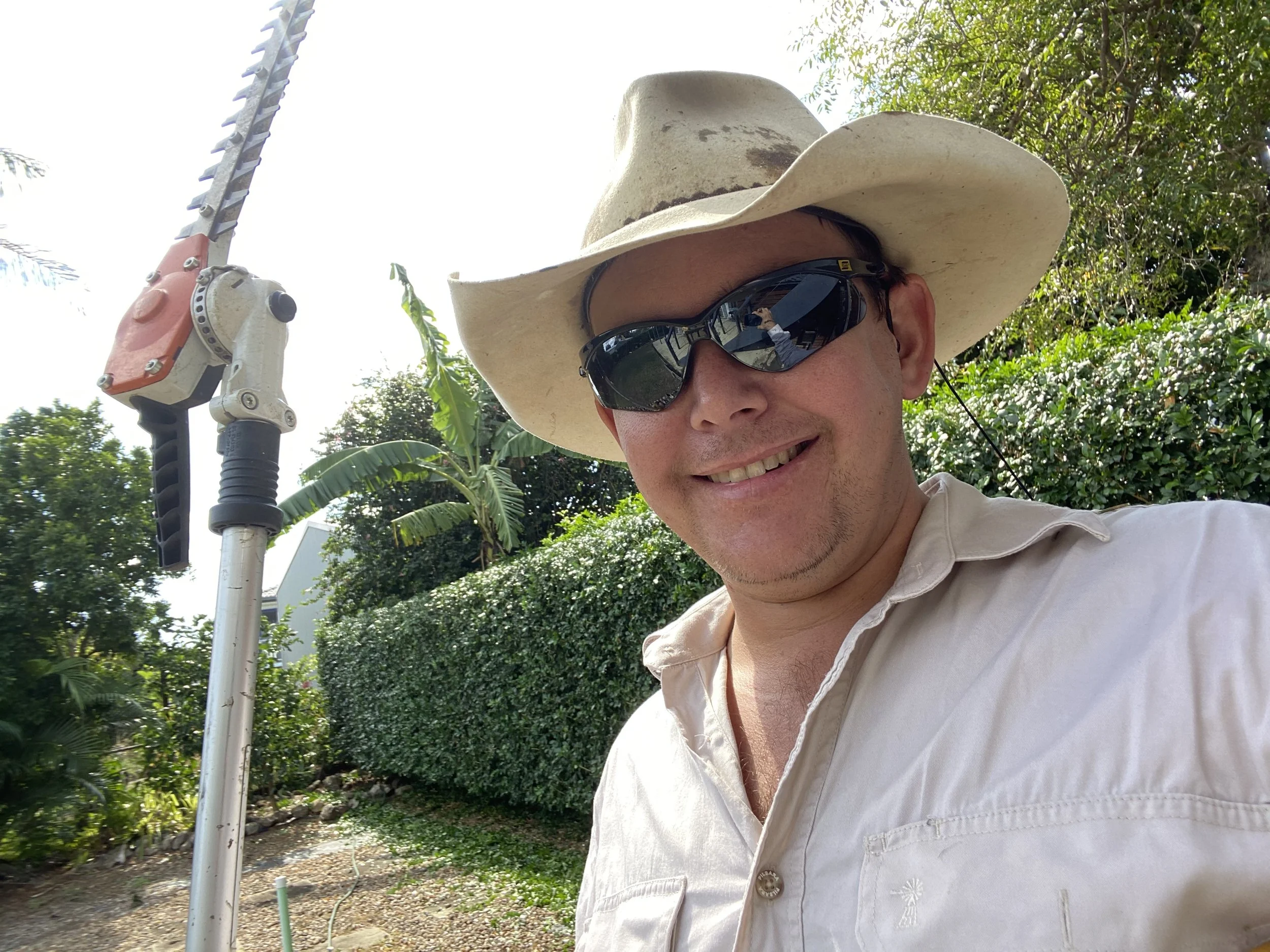 A man with a big sunhat and sunglasses smiling outdoors surrounded by green trees and plants.