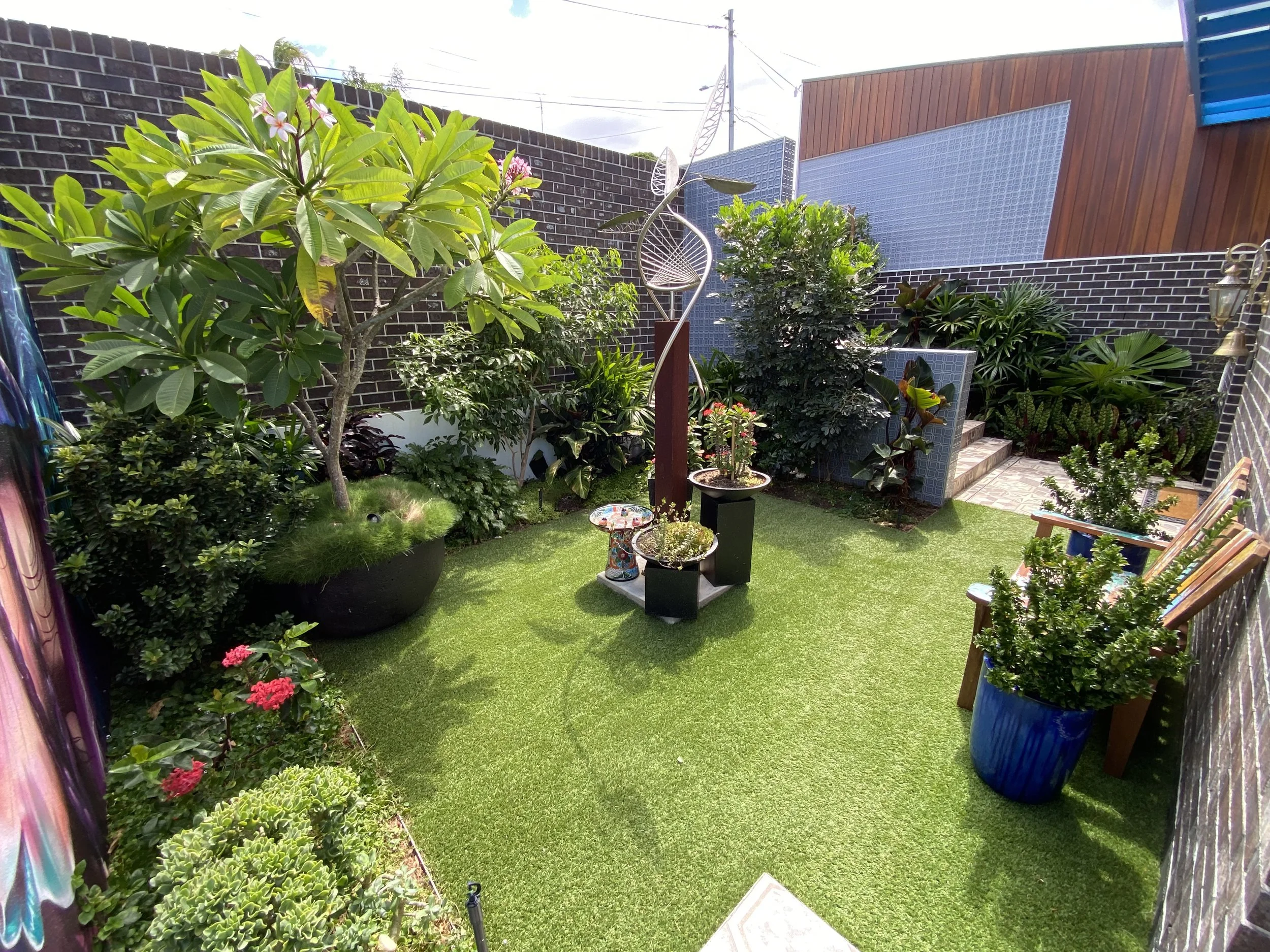 A small, colorful backyard garden with green grass, potted plants, trees, and outdoor furniture, enclosed by dark brick walls with decorative panels.