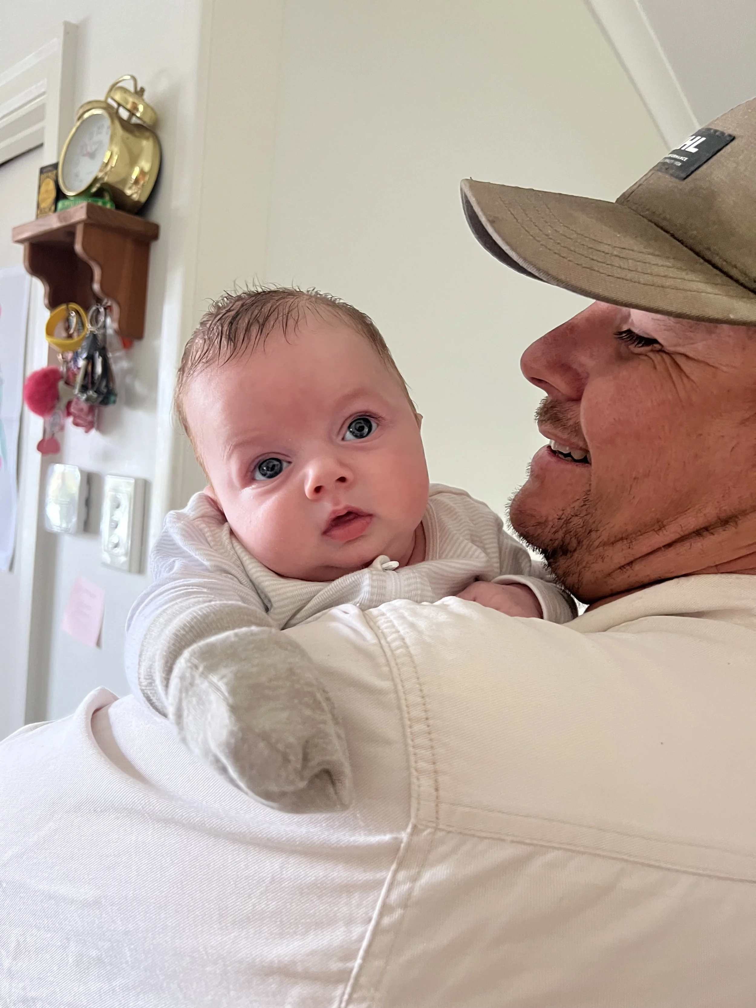 A man with a tan baseball cap holding a baby with big blue eyes and light brown hair, both smiling in a room with a white wall and a wooden key holder with keys and a clock in the background.