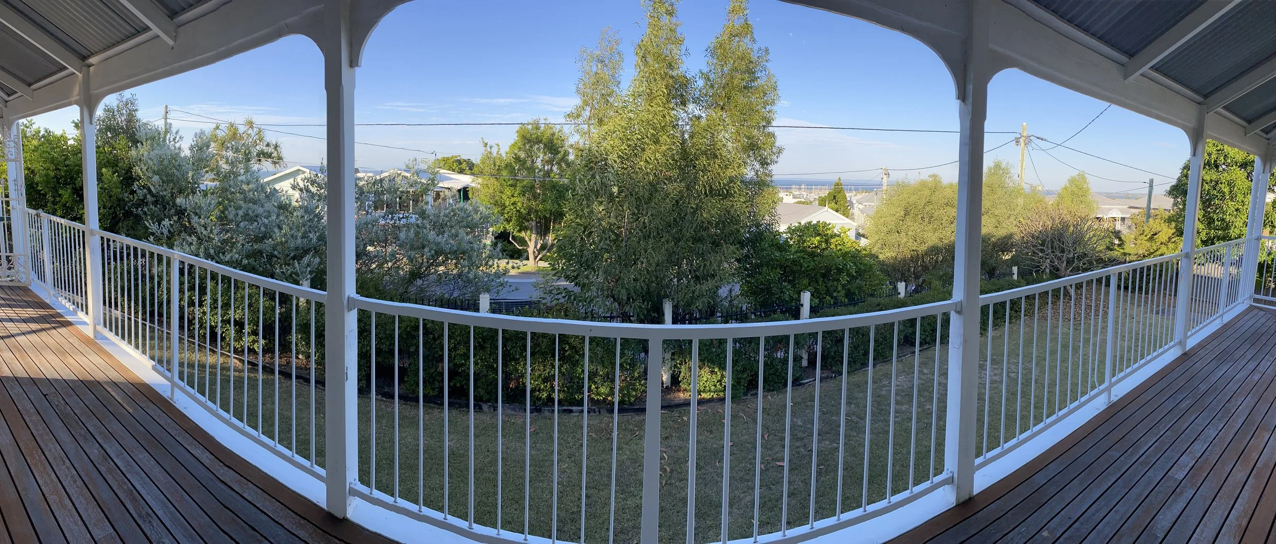 Panoramic view from a covered balcony with wooden flooring and white railing, overlooking a backyard with greenery and trees, with houses and power lines in the distance under a clear blue sky.