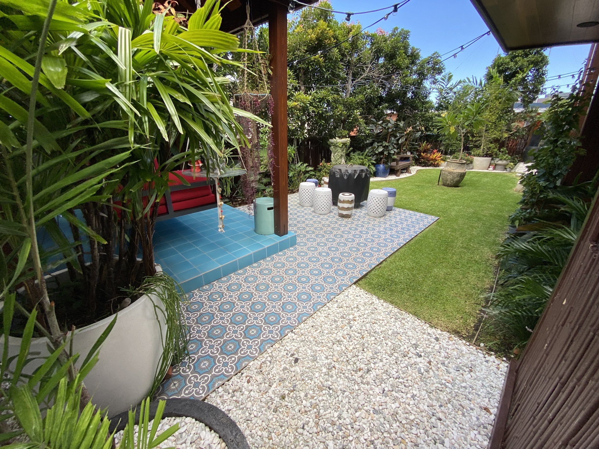 A backyard patio area with patterned tiles, surrounded by greenery including potted plants, trees, a stone pathway, and grass, under a blue sky with some string lights overhead.