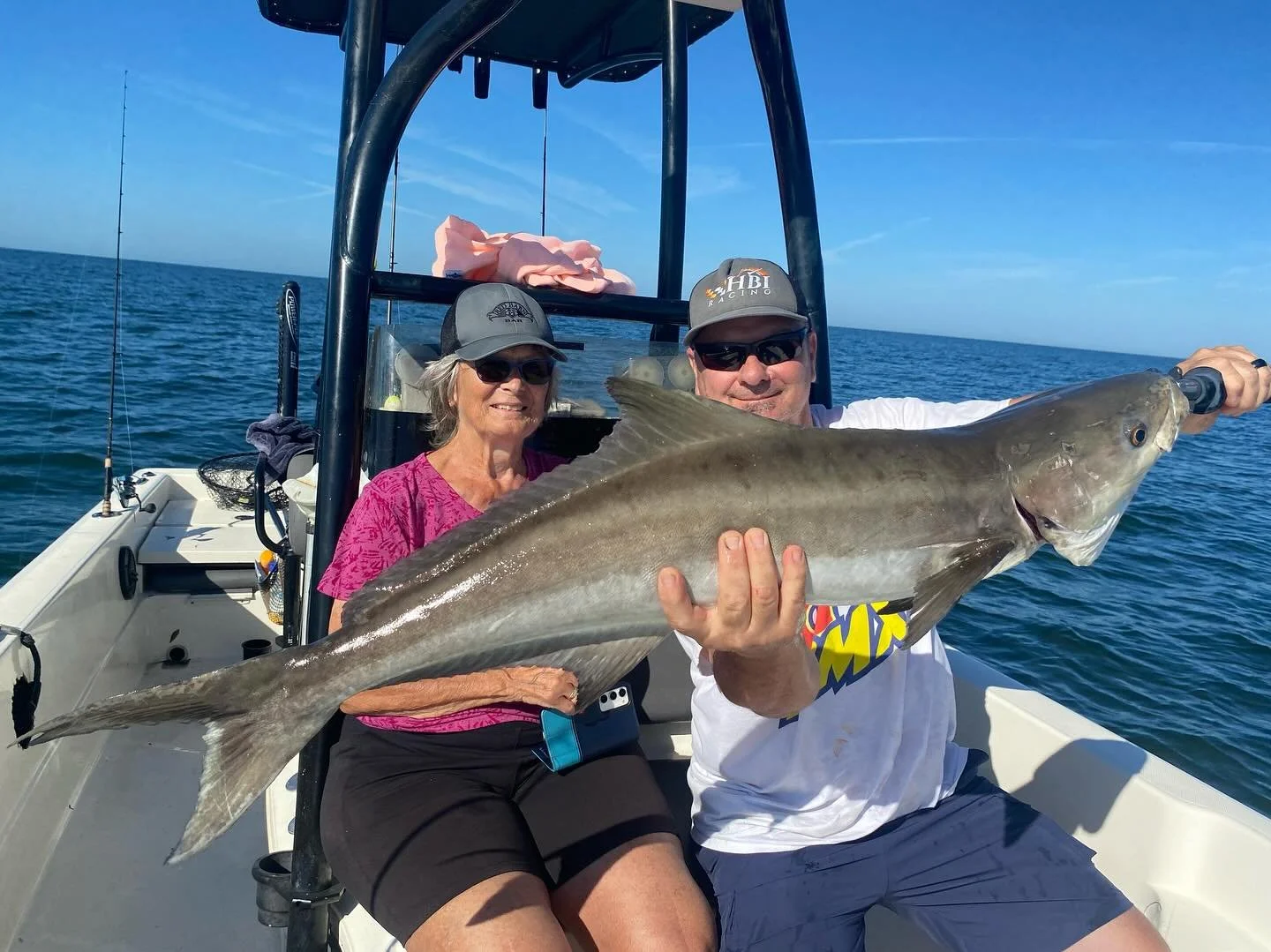 A man and woman on a boat holding a large fish, with the open ocean in the background. The man is wearing sunglasses and a cap, smiling and holding the fish with both hands, while the woman sits next to him, also smiling and wearing sunglasses and a