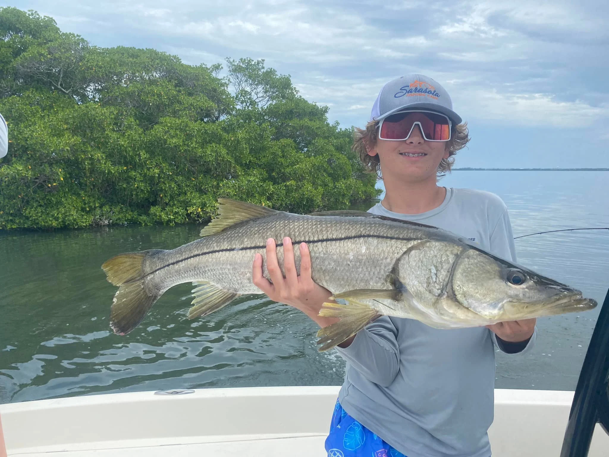 Young boy wearing sunglasses and a cap holding a large fish on a boat with water and green trees in the background.